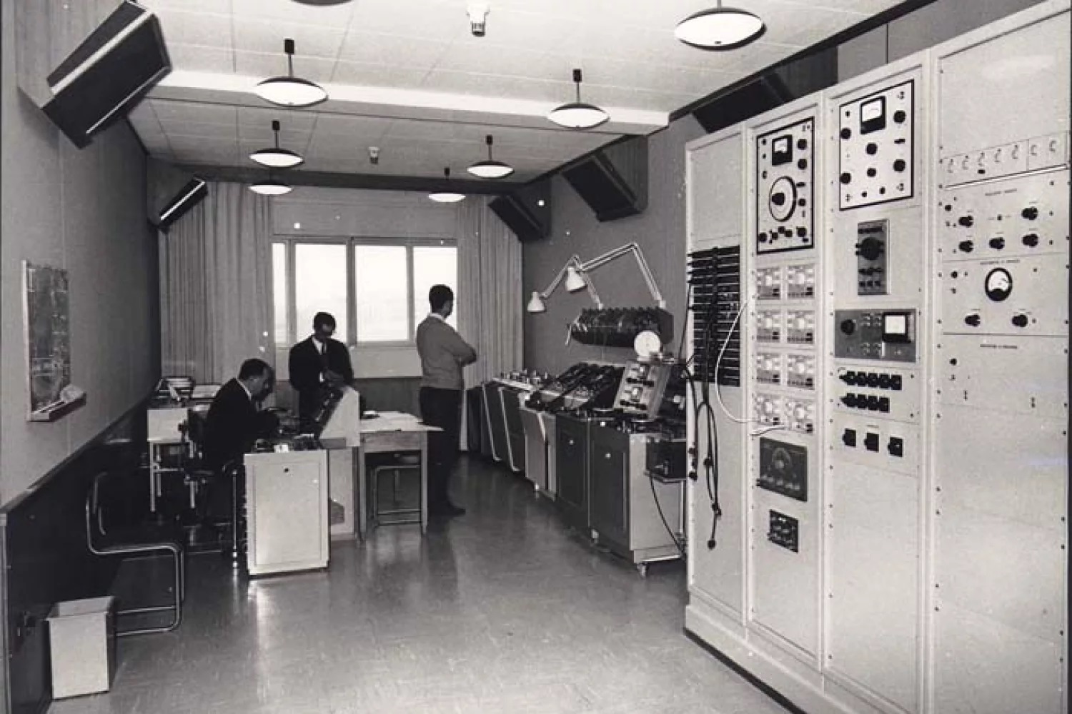 Black and white image of a vintage computer room with large electronic panels. Three men work at desks and control units.