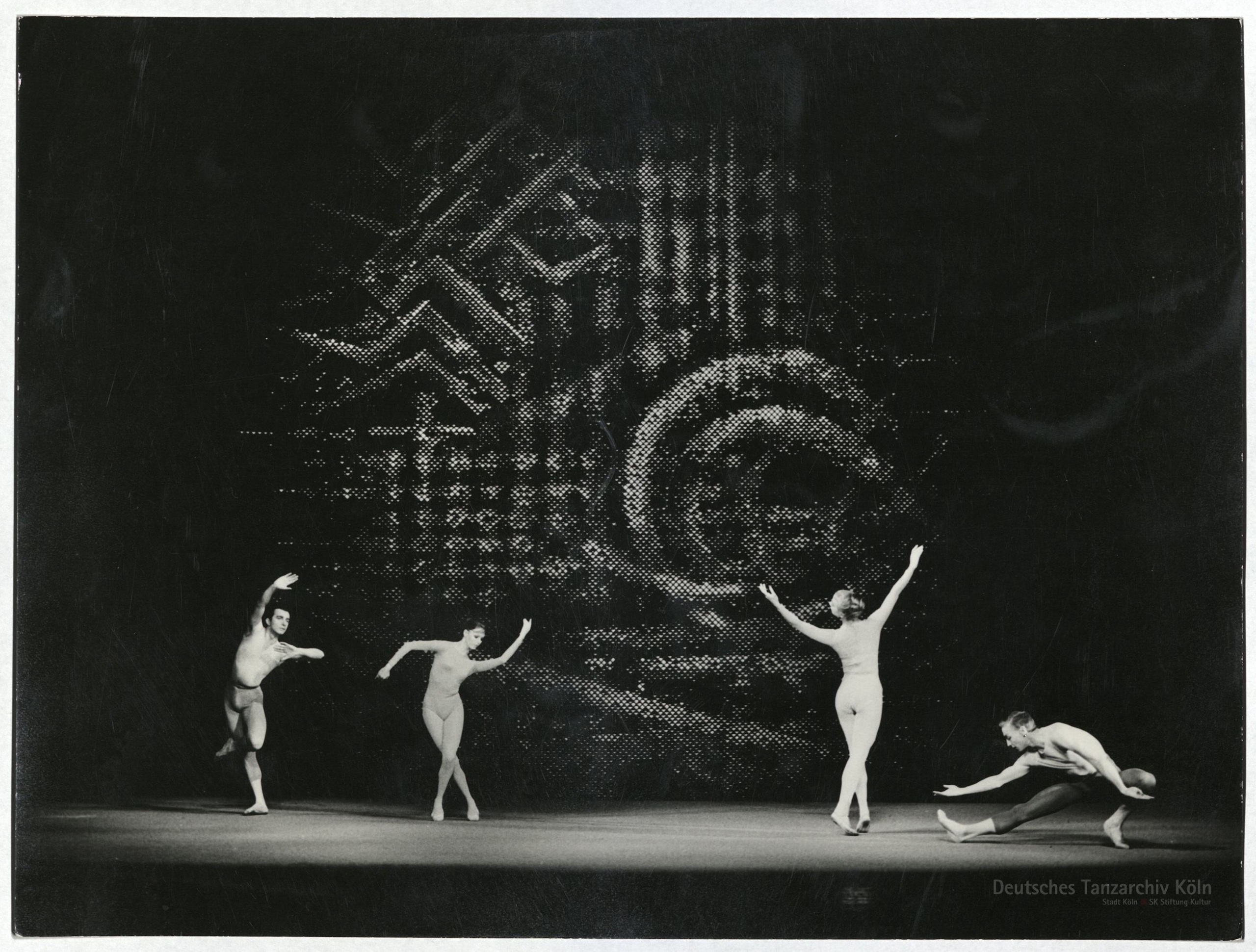 Four ballet dancers perform on a dark stage. The backdrop features abstract, large-scale patterns.