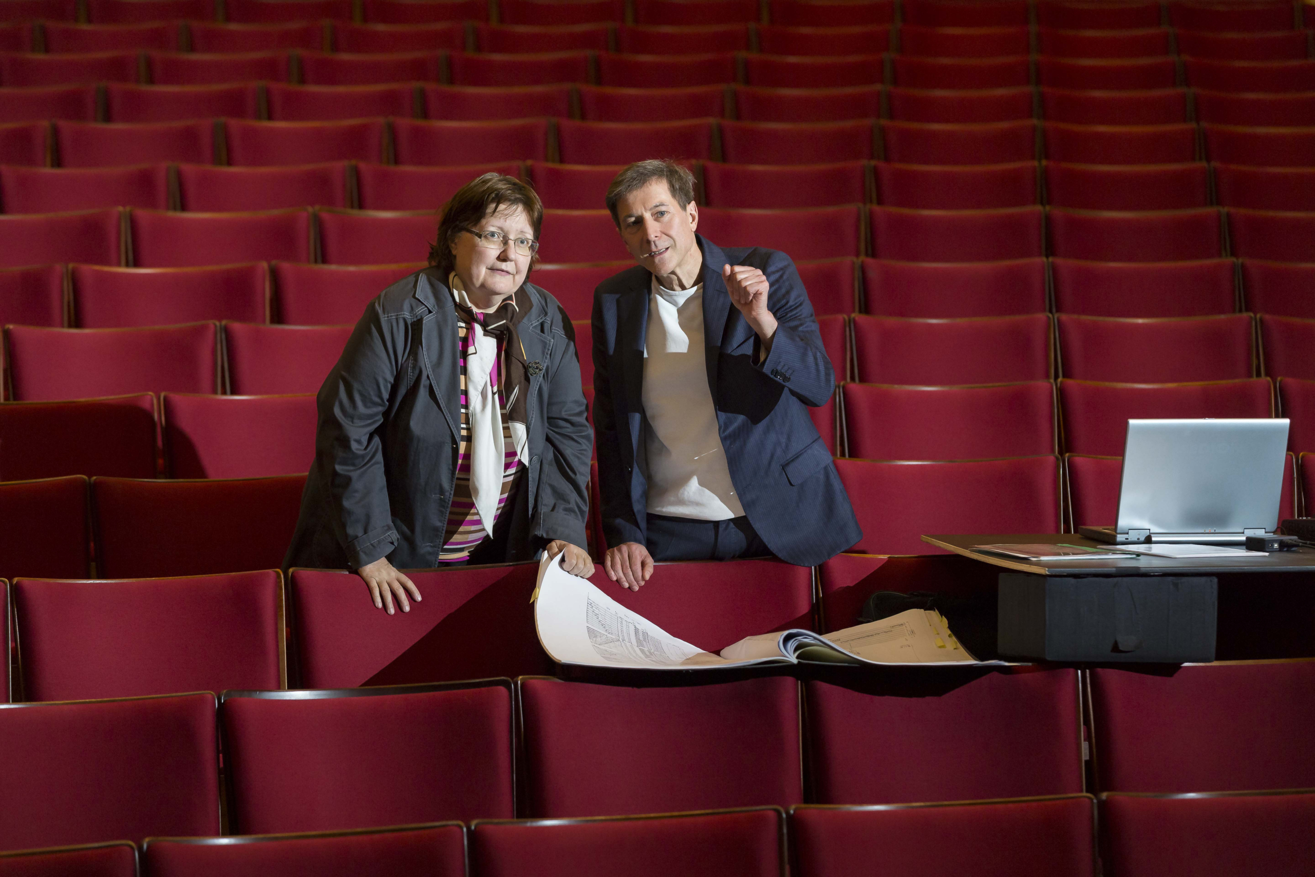 Two people are standing in an empty theatre auditorium with red seats, talking and going through documents while looking at the stage. Next to them is an open laptop.