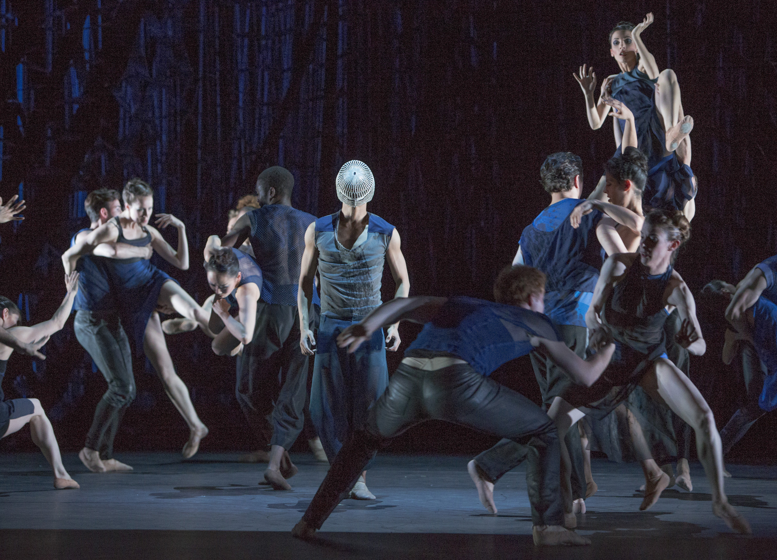 A group of dancers in modern costumes perform dynamically under dim lighting. A central dancer stands out with a white headpiece.
