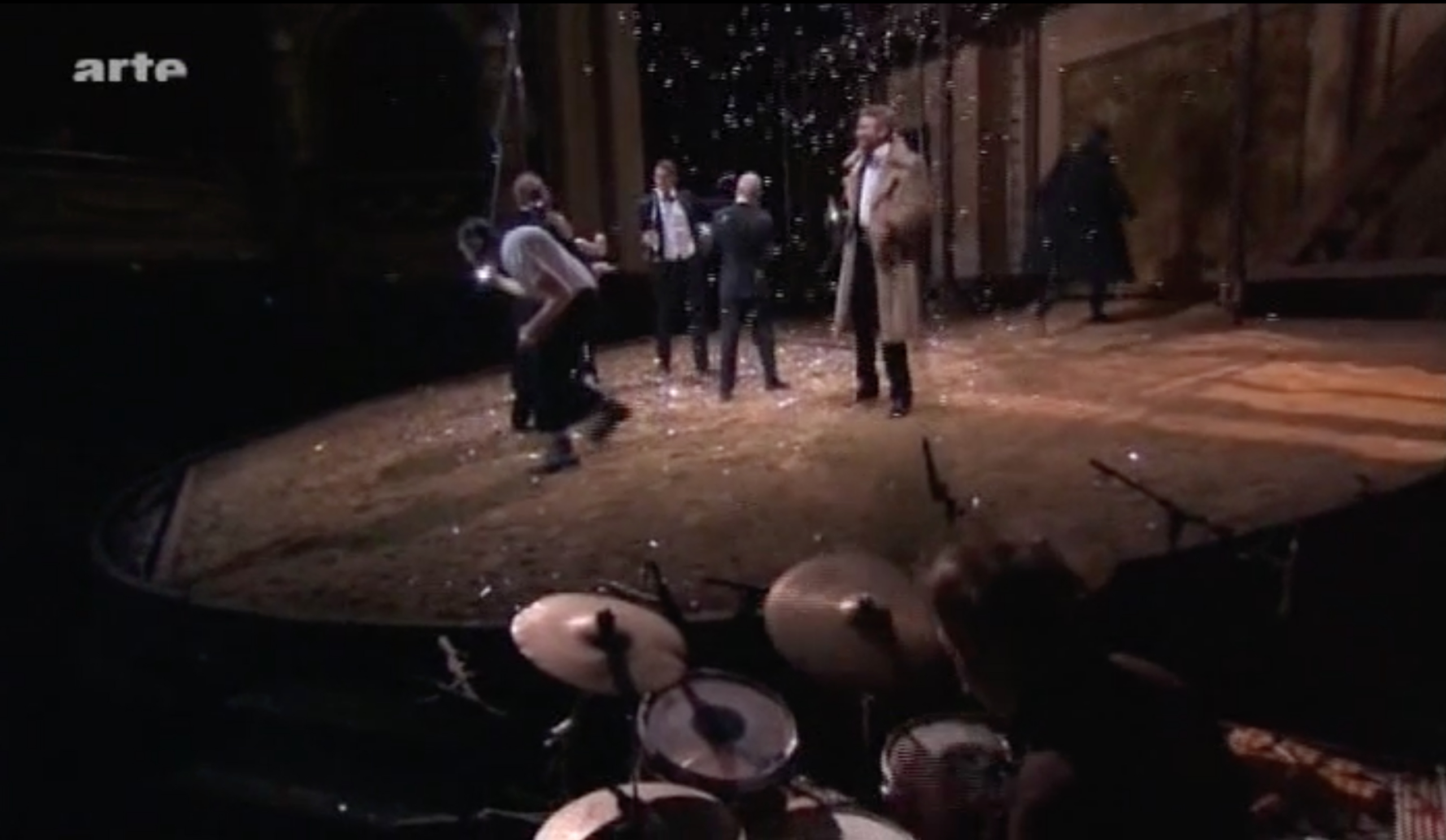 A video still shows a stage with six performers behaving animatedly and a drummer playing in the foreground.
