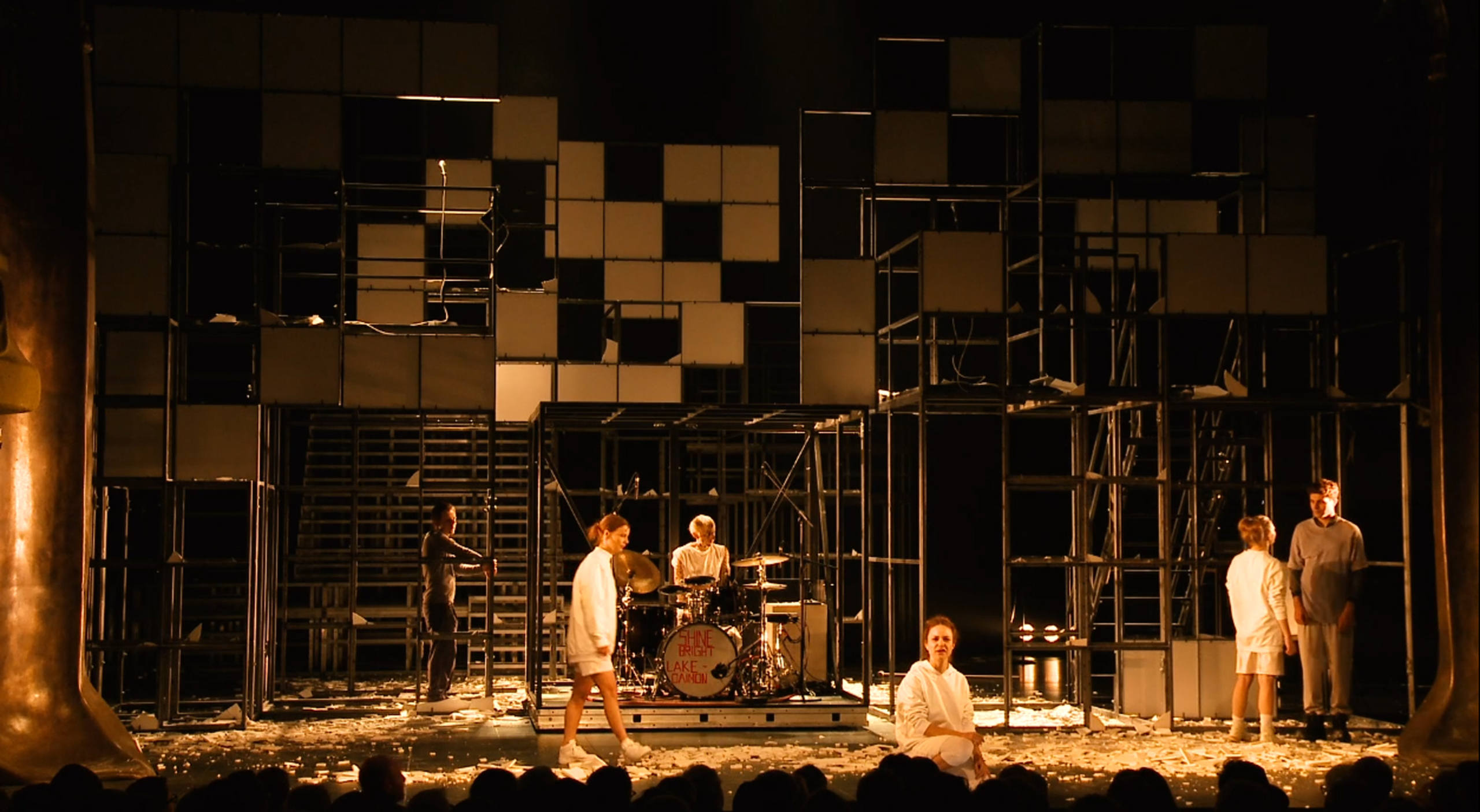 On a stage with a modern geometric backdrop, six performers in white are in view, one of whom is playing the drums. The stage is lit in a warm, orange-coloured tone.
