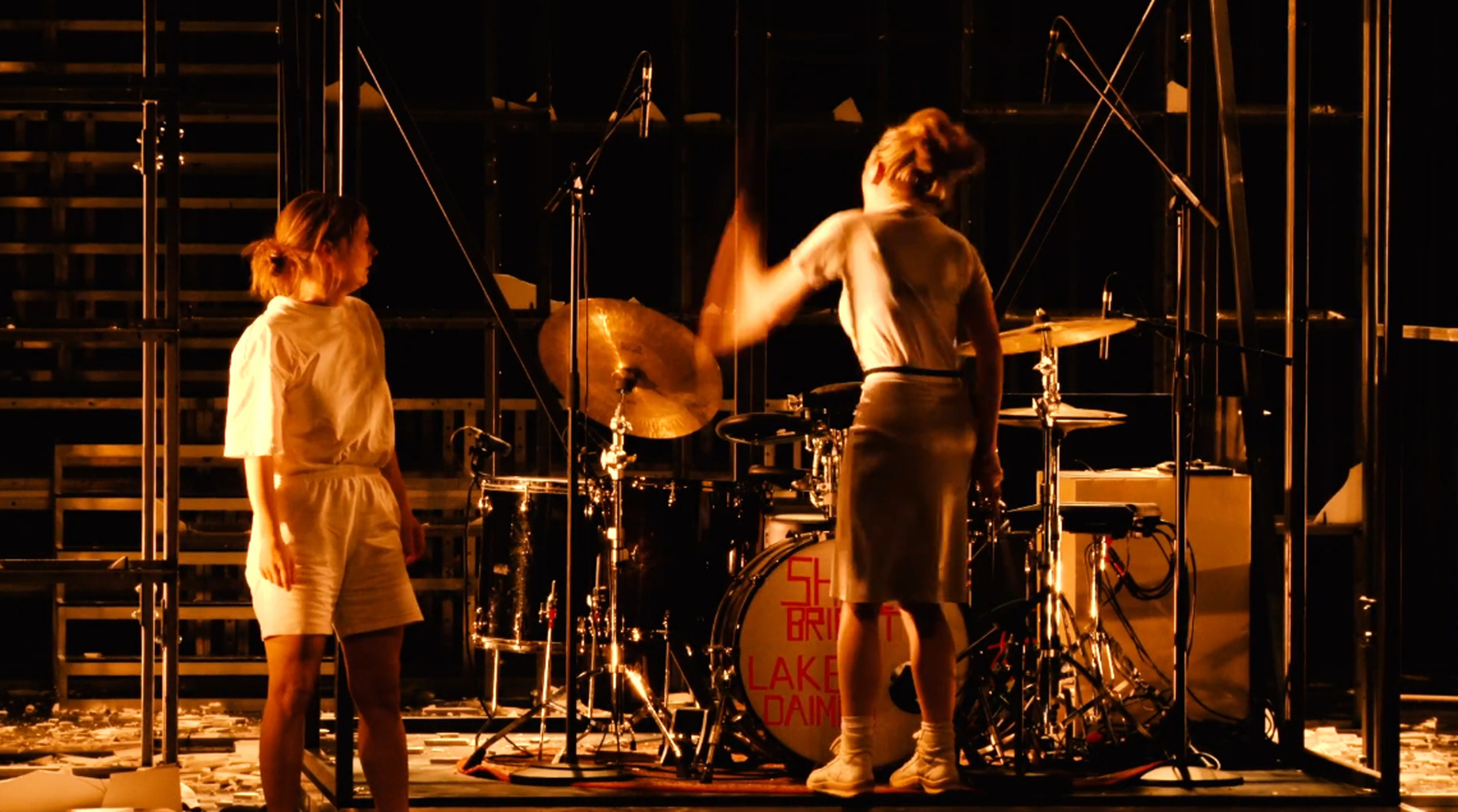 Two performers are standing on a stage lit in a warm orange tone. One of them plays the drums energetically while the other watches.