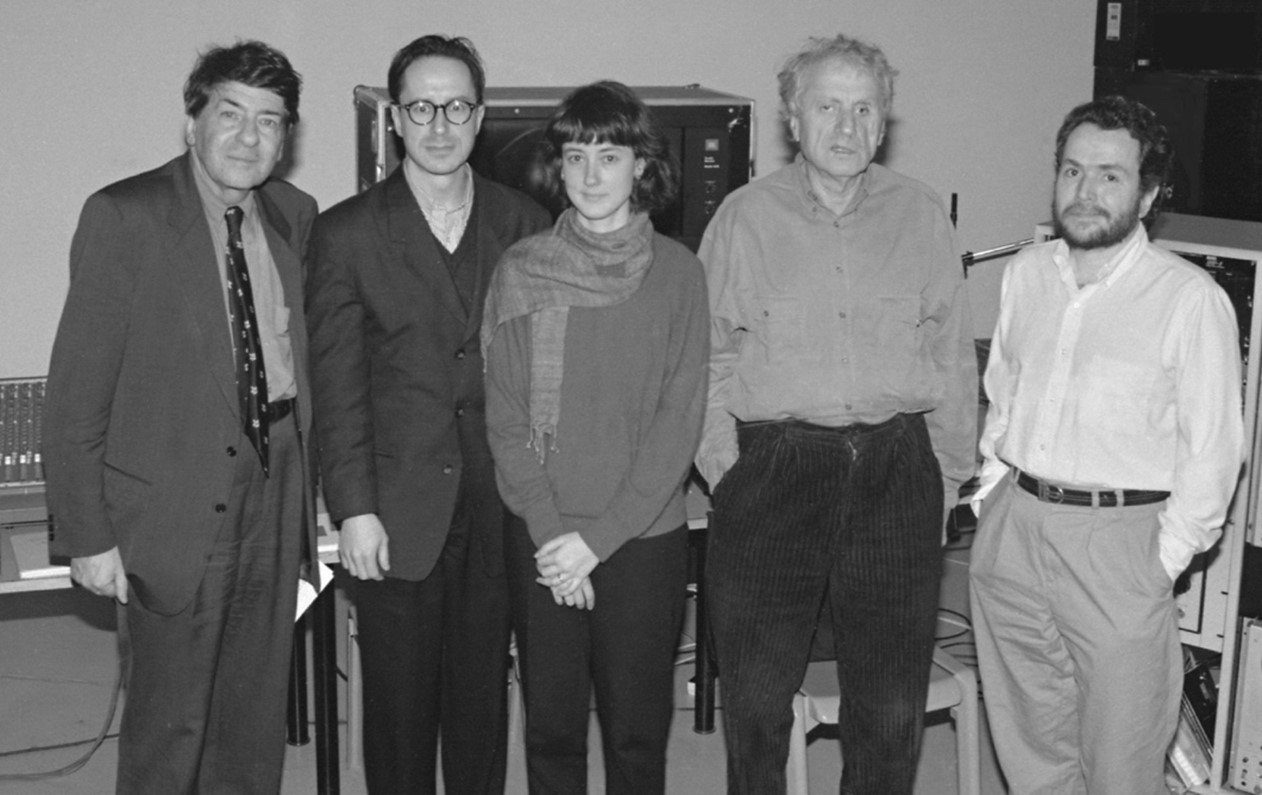 Five people stand closely together in a black and white photo, dressed in formal and casual attire. The background shows audio equipment, suggesting a studio setting. Their expressions are neutral.