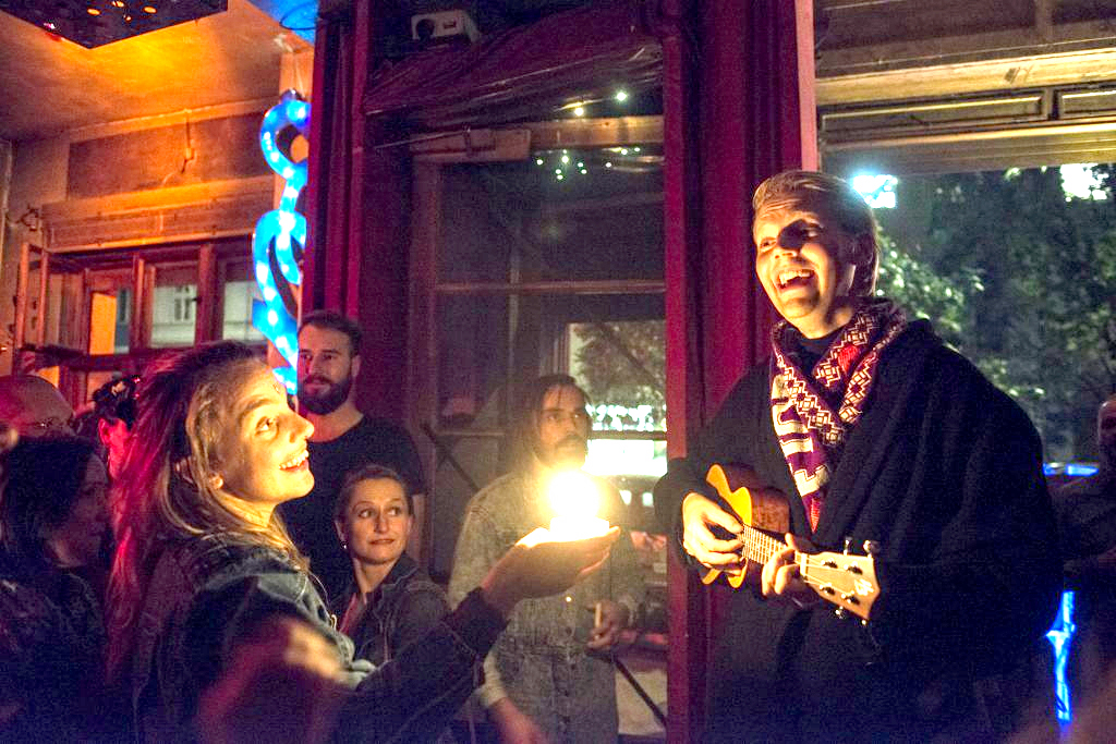 A smiling musician plays a small guitar inside a lively bar with warm red lighting while another person holds up a glowing candle toward them. Other people are gathered around.