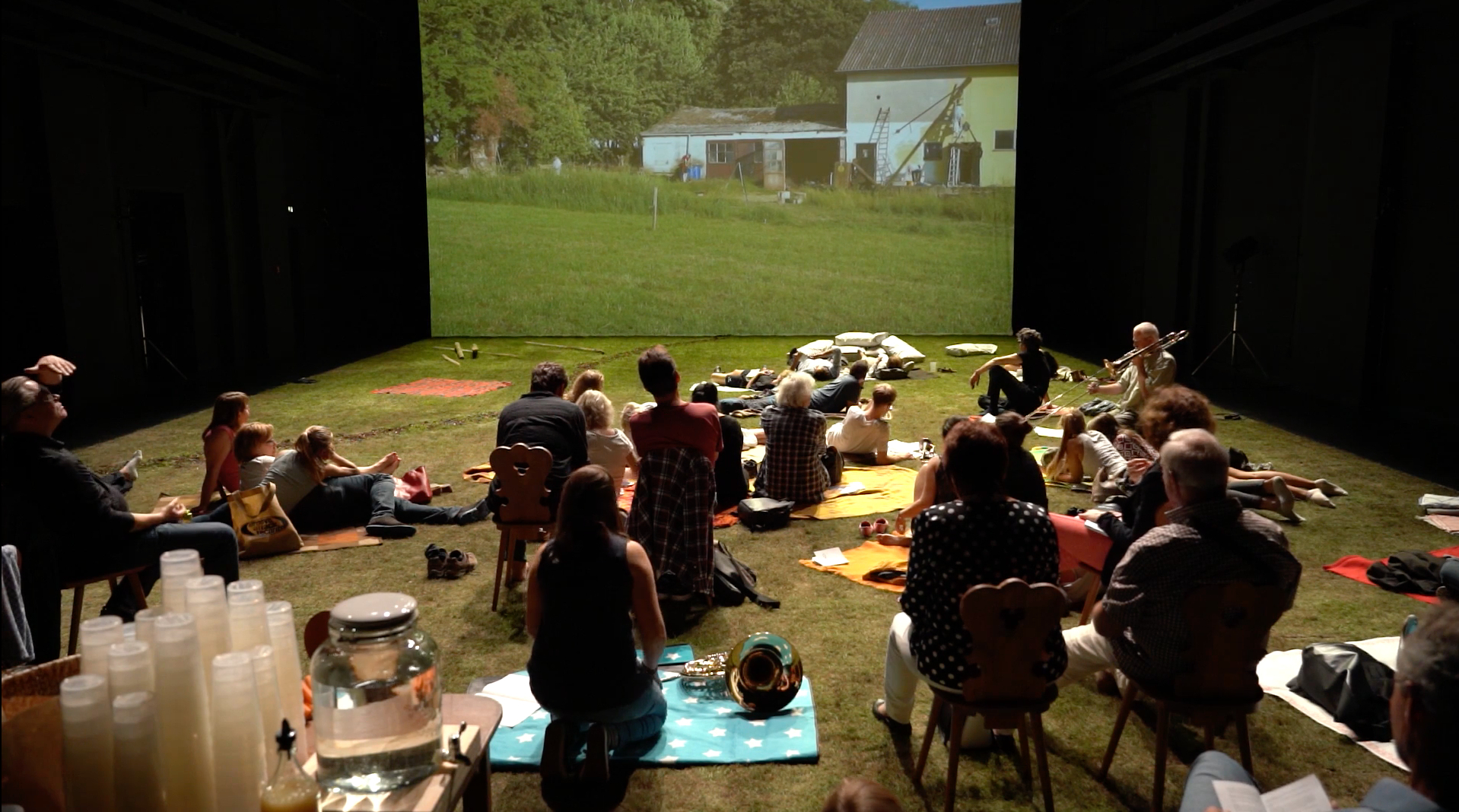 In a dark space, people gather on a grassy floor in front of a large screen and watch a rural scene featuring grass.