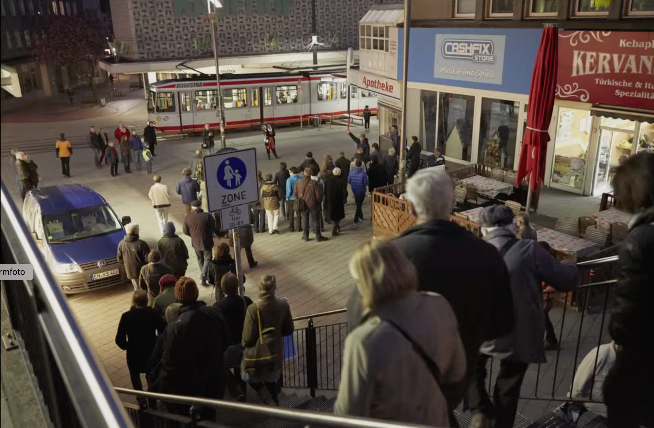 A man in formal attire stands in front of glass windows and speaks to an audience. The windows look out onto an urban street.