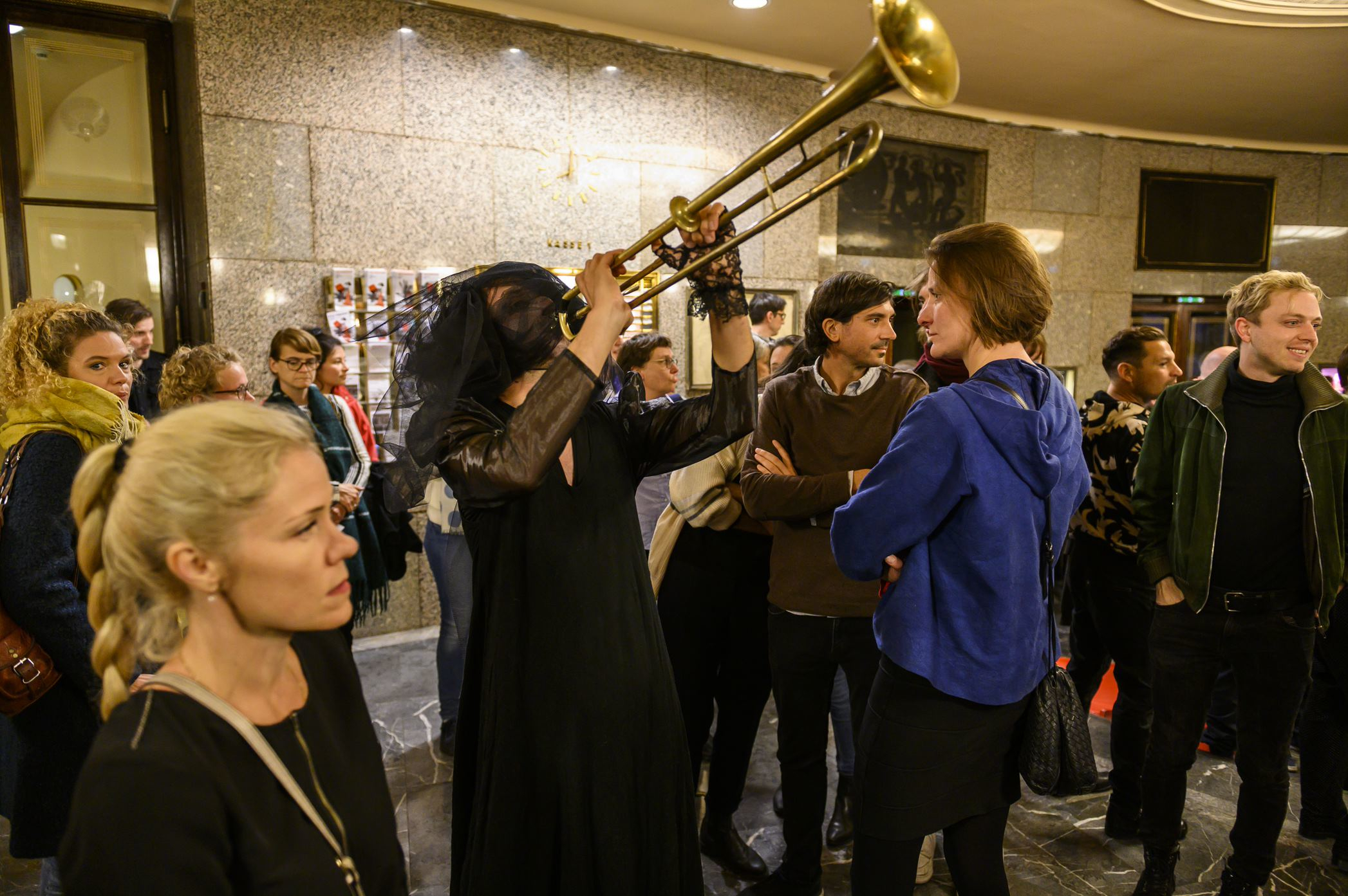 A person dressed in black with a veil plays a trombone in a busy box office foyer of a theatre.