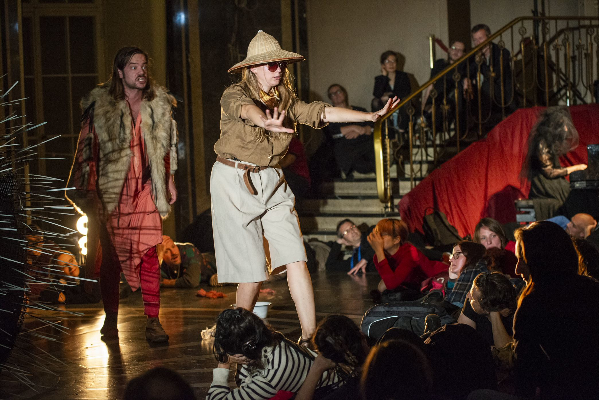 Two performers stand in the spotlight, one is wearing a fur coat, the other is wearing safari clothing and gesticulates. The audience sits on the stairs and lies on the floor.