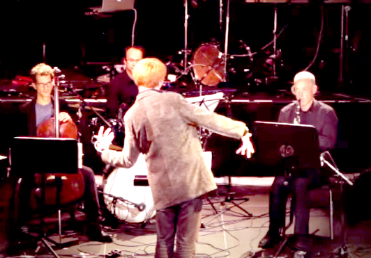 A conductor passionately leads a trio of seated musicians, framed by a dimly lit stage with a drum set behind them.