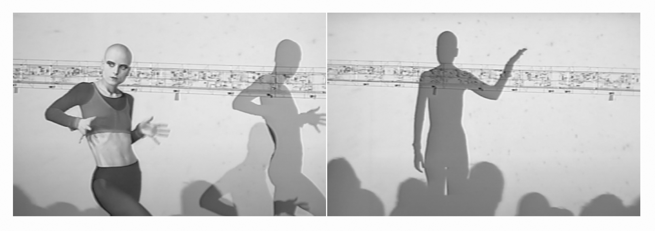 Black and white photo of a performer with shaved head, wearing a fitted top, dancing energetically against a backdrop with shadow silhouettes.