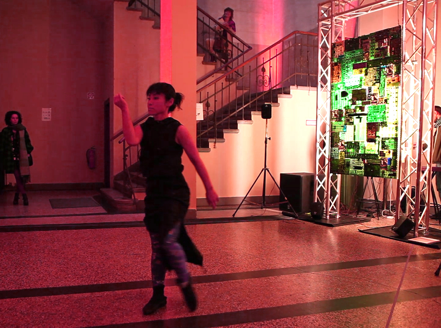 A dancer in a dark outfit performs gracefully in a dimly lit room with a large electronic display emitting green light. A staircase and onlookers are in the background.