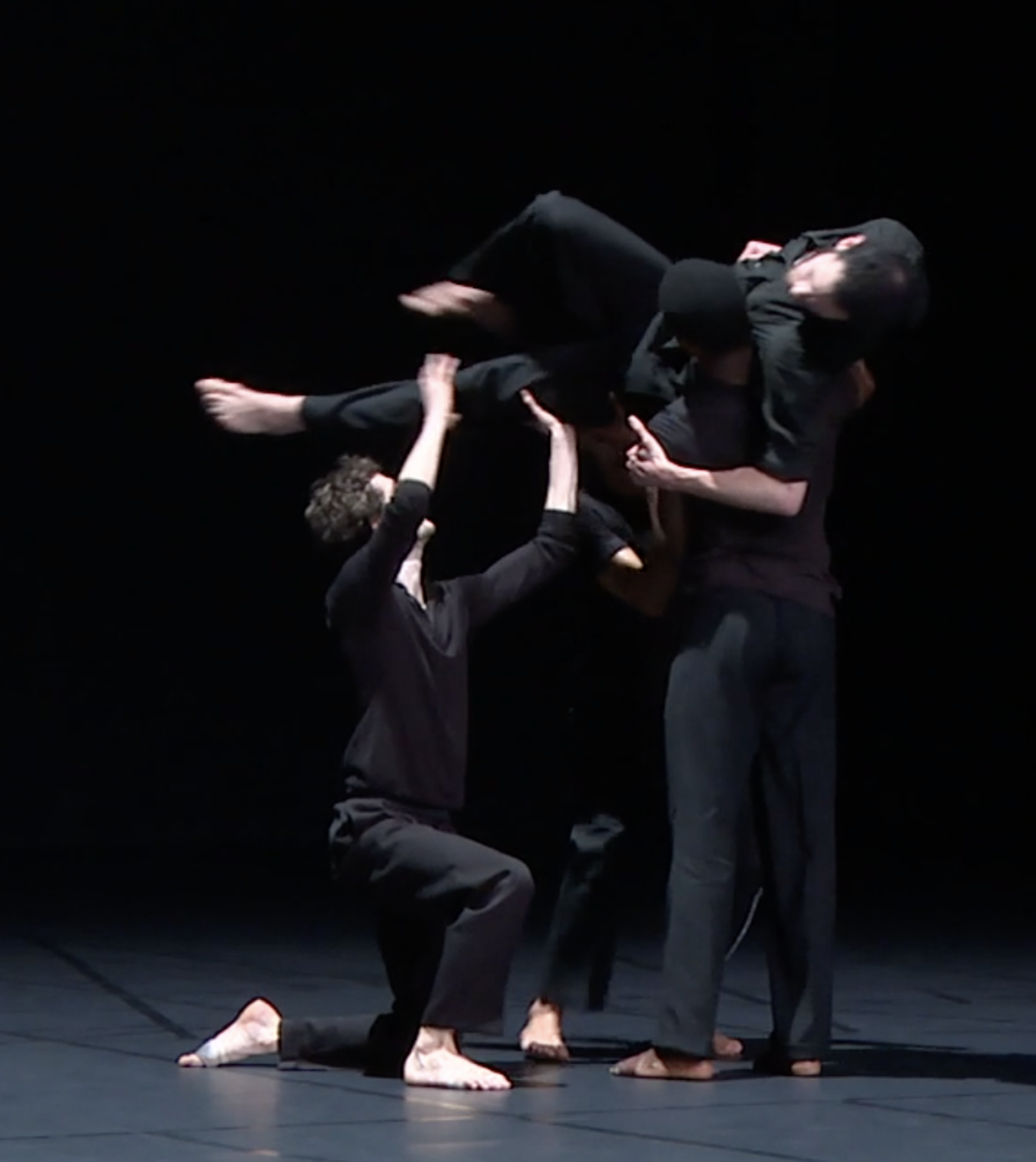 Four dancers in black perform a dynamic routine. Two dancers lift a third horizontally, while the fourth kneels, supporting.