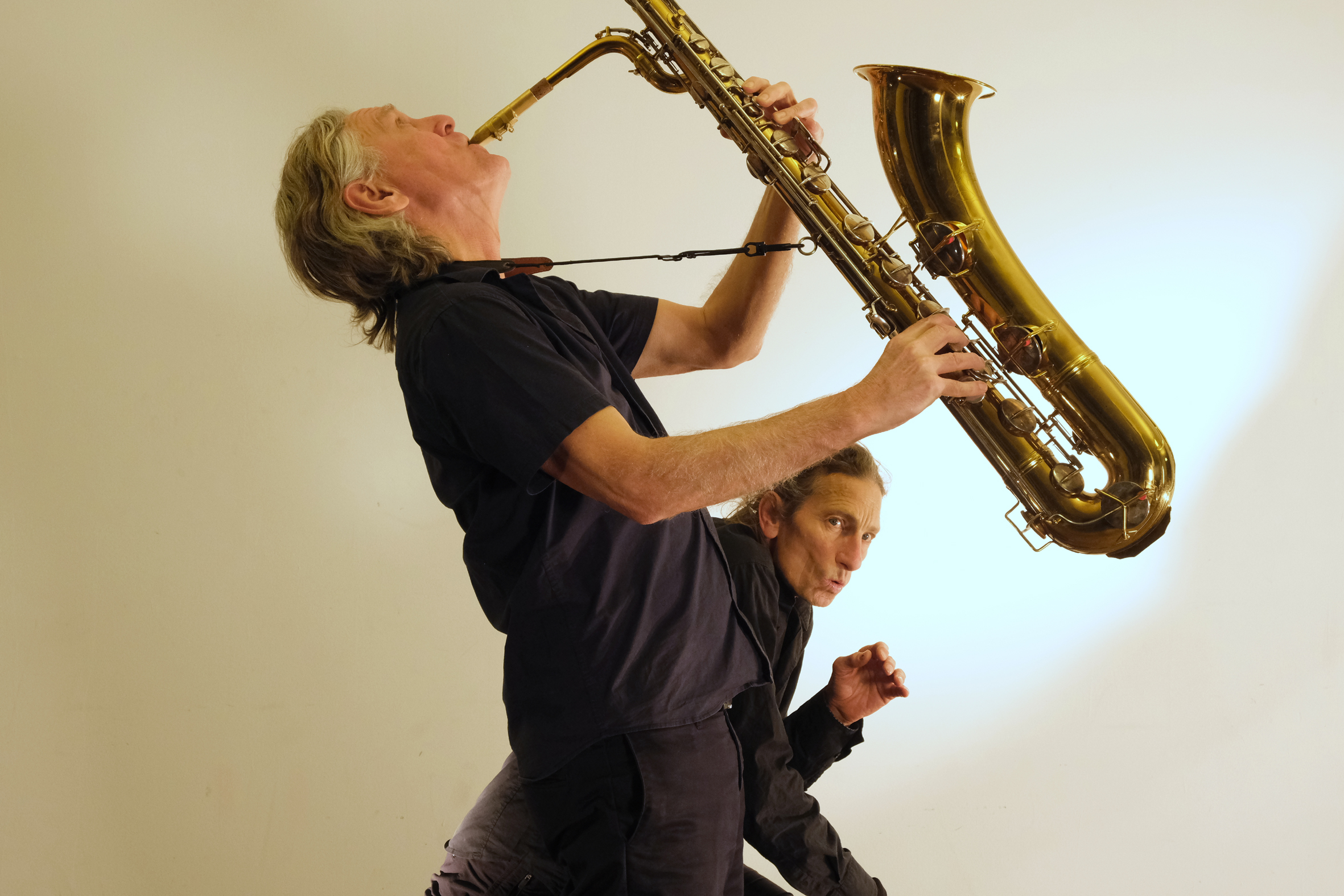 Two musicians perform dynamically against a light backdrop. One plays a large saxophone with enthusiasm, while the other gestures expressively.