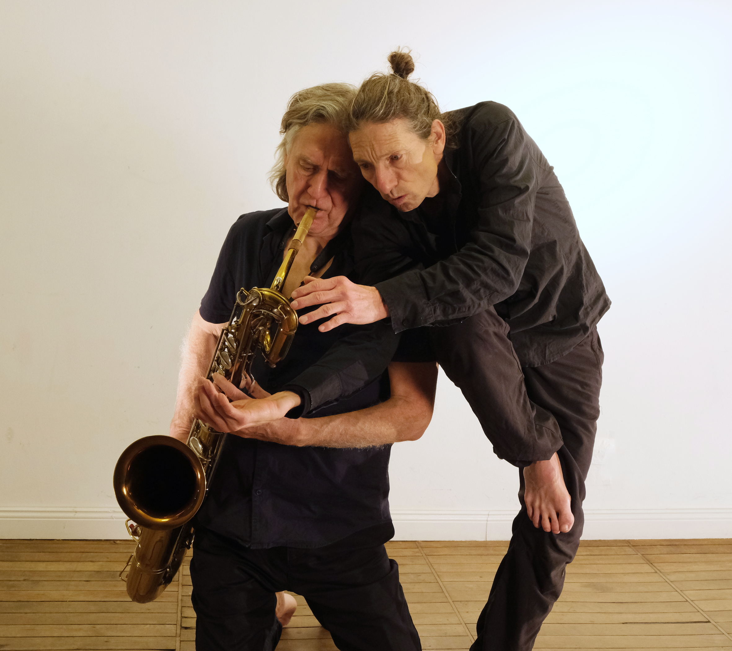 Two men in black clothing engage in an expressive performance. One plays a saxophone while the other leans on him.