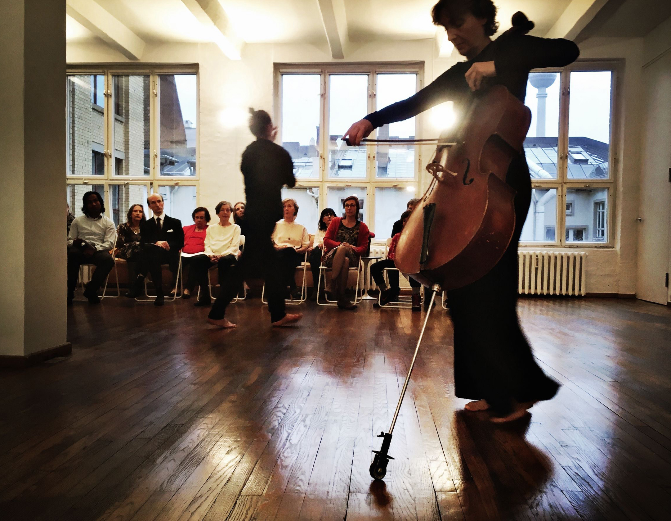 A cellist in dark clothing performs barefoot in a spacious room, conveying focus and passion. An attentive seated audience observes. Soft lighting creates a cozy ambiance.