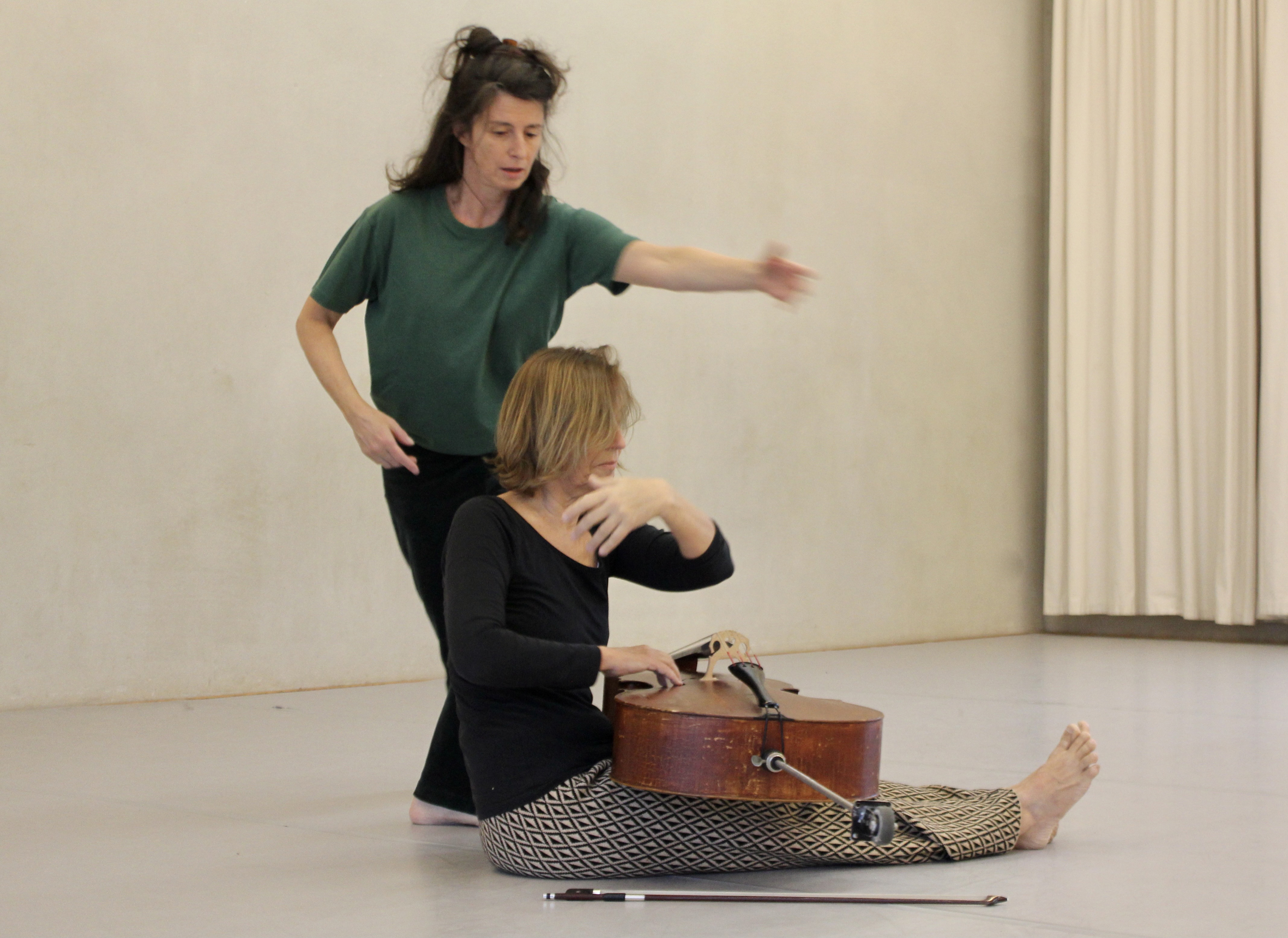Two women in a serene dance studio. One sits cross-legged, playing a cello, while the other gestures expressively, conveying artistic collaboration.