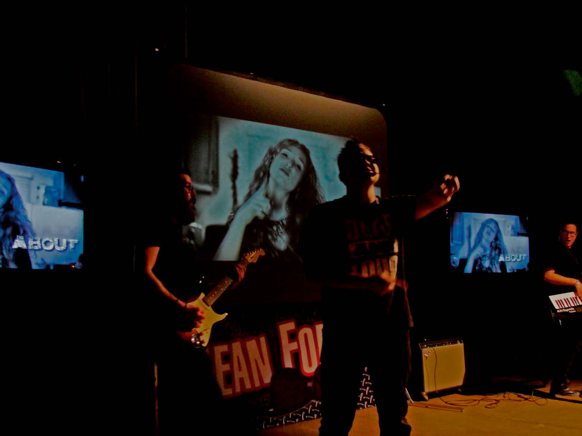 Three people on a dark stage, one with an electric guitar, one lifting the left arm chest-high, one with a keytar. There are three identical pictures in the background, showing a person making a gesture with the left hand and a lettering saying 