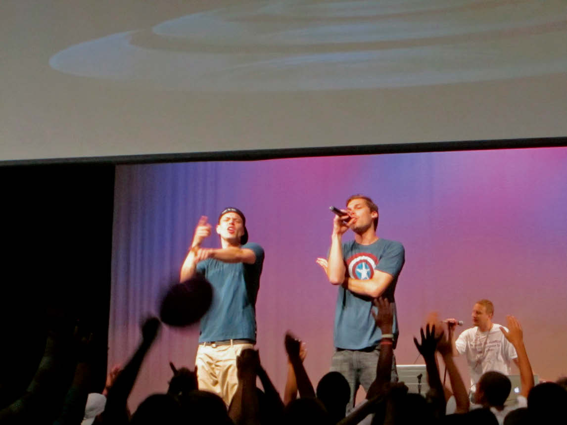 Three people on a stage in front of an audience raising many hands, two with microphones, one making gestures.