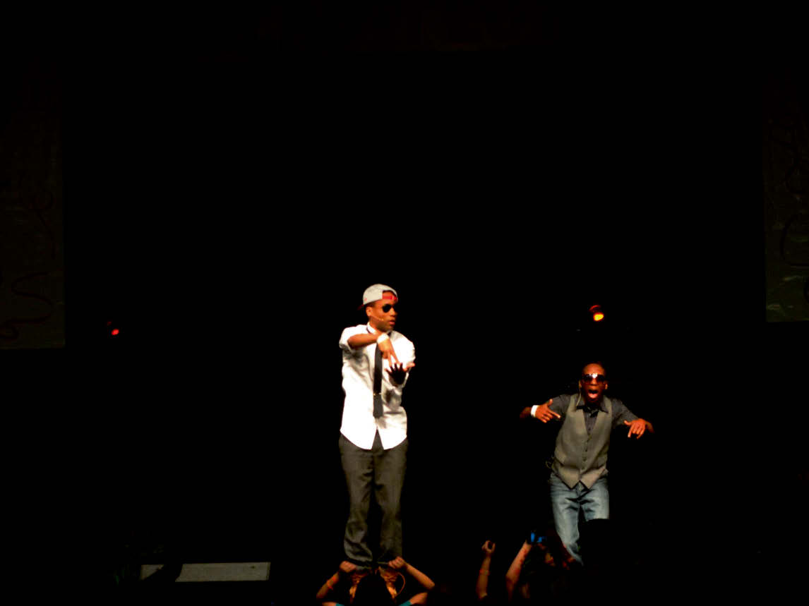 Two people on a stage making gestures in front of an audience with some raised hands.
