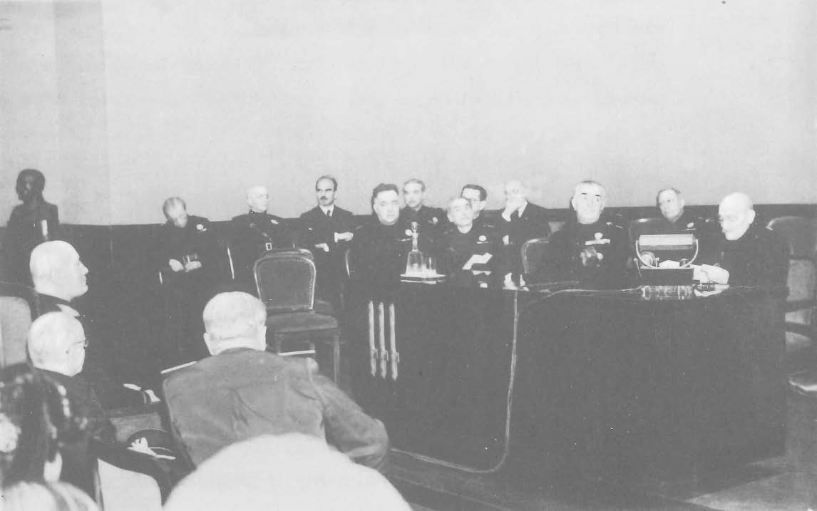 Four people sitting behind a desk on a stage with seven people sitting in their backs and an audience in front of them.