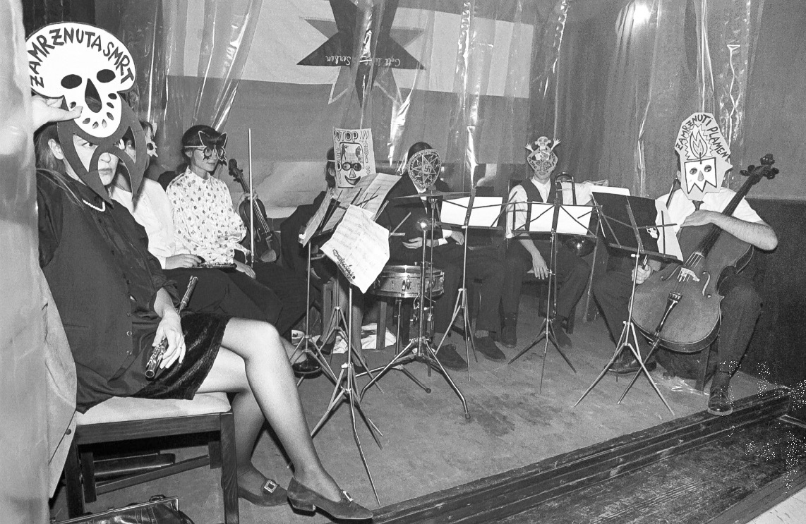 Seven people sitting on a stage with musical instruments, wearing paper cut-out and painted face masks.