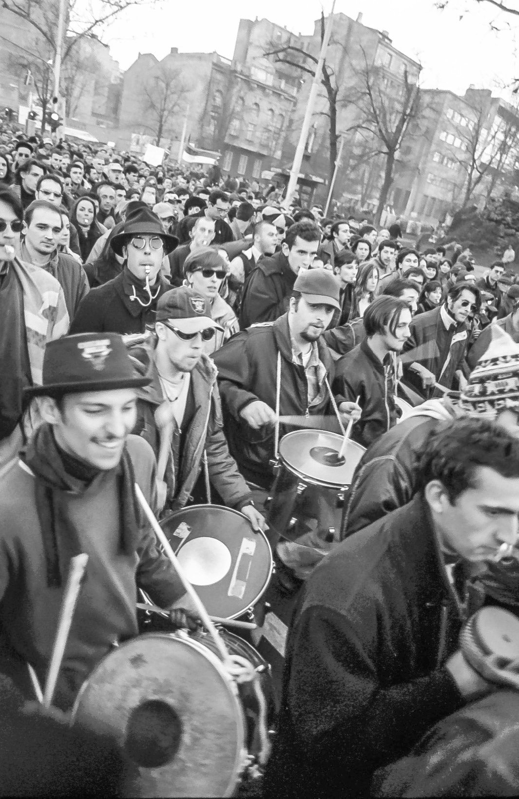 A large crowd of people, some playing percussion instruments and whistles in the foreground, in the background appartment buildings of varied dimensions and street-lining trees without leaves.