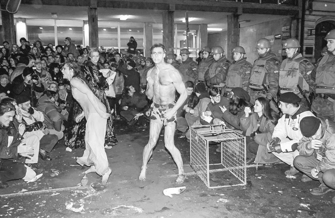 Two people performing lightly dressed in front of a building with a cage that could fit a middle-sized dog, surrounded by an audience and policemen.
