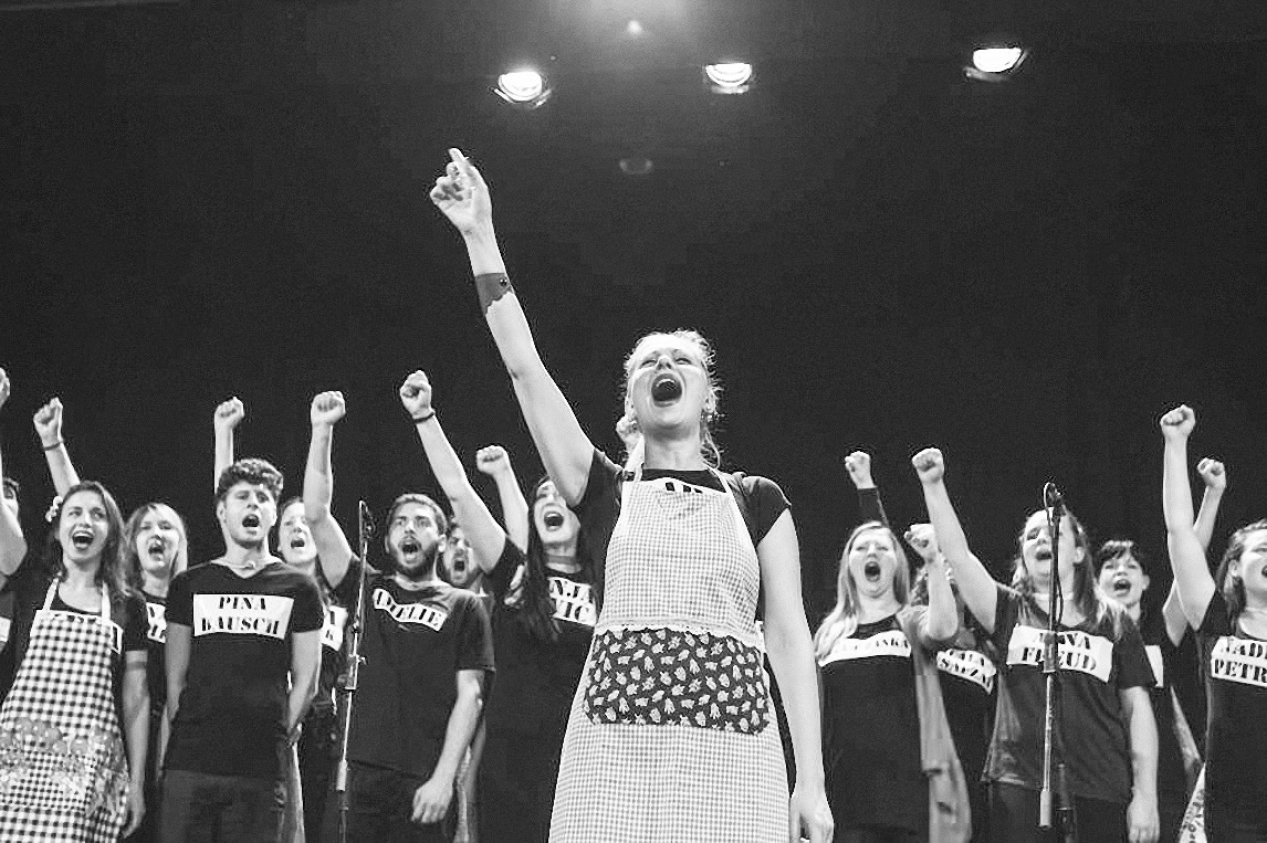 15 people, most of them raising a fist, with open mouthes and similar shirts, one up front wearing an apron.