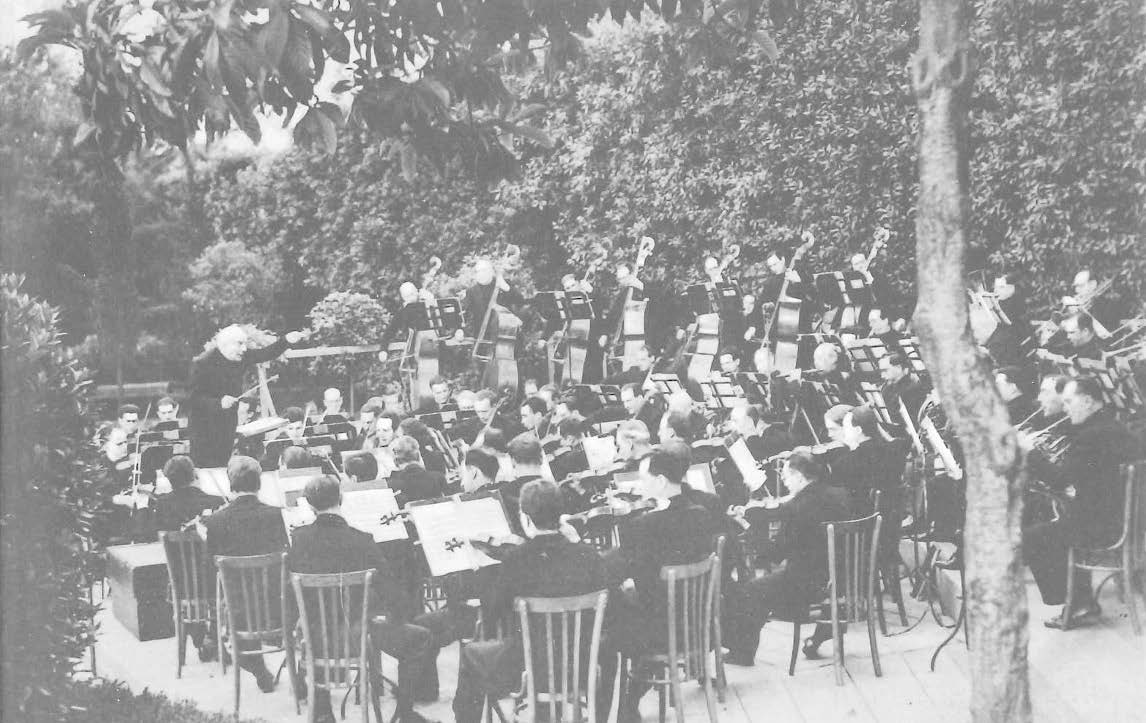 Orchestra playing in a garden, seen from behind the strings, facing the conductor.