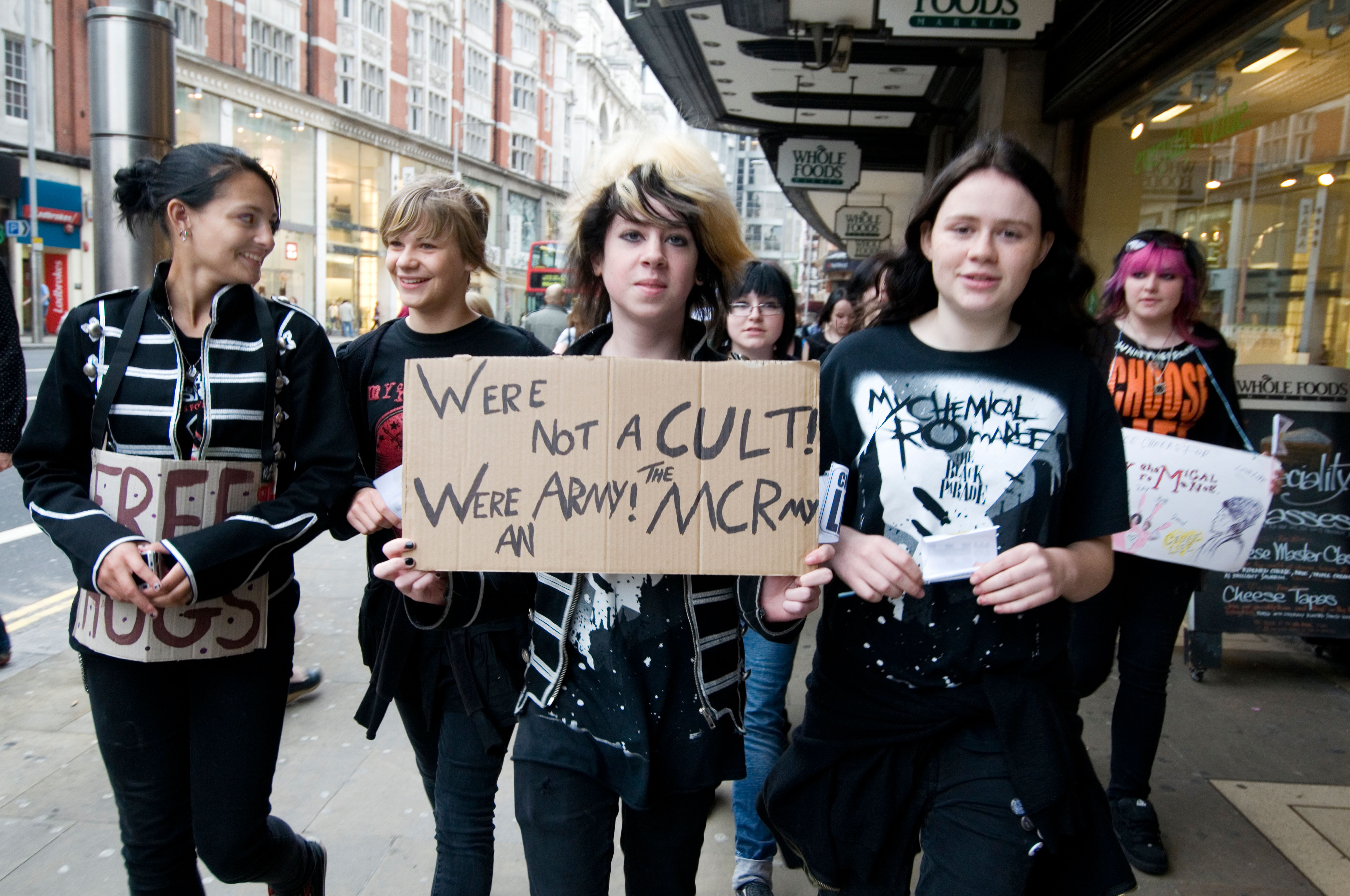 Young, smiling female EMO fans protesting. They are holding a cardboard sign saying »Were not a cult! Were an army! The MCRmy«.