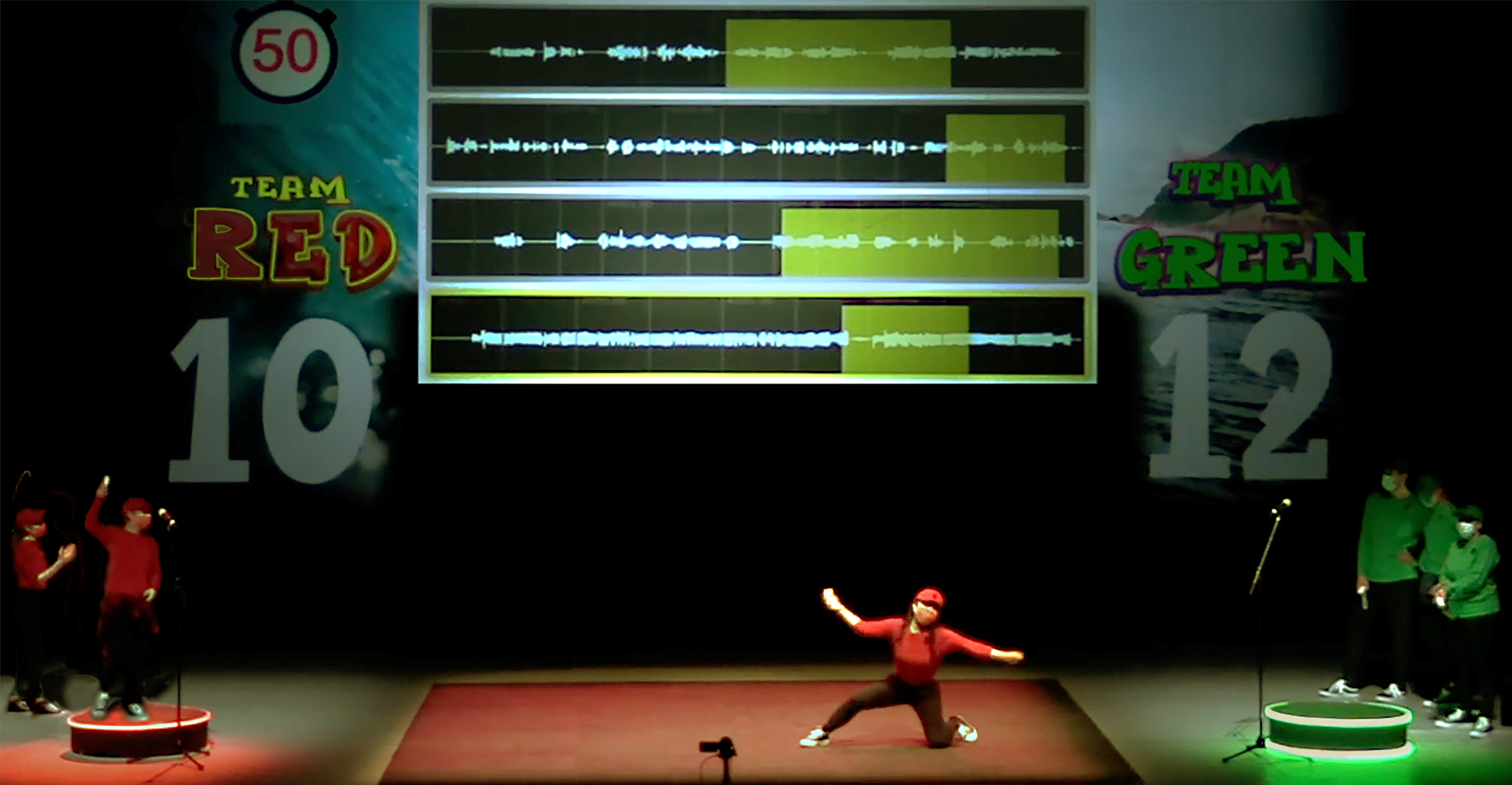The image shows a team in red and a team in green compete on a stage with a large screen displaying their scores and audio waveforms. A performer in red is kneeling in the center, and a timer shows “50”.