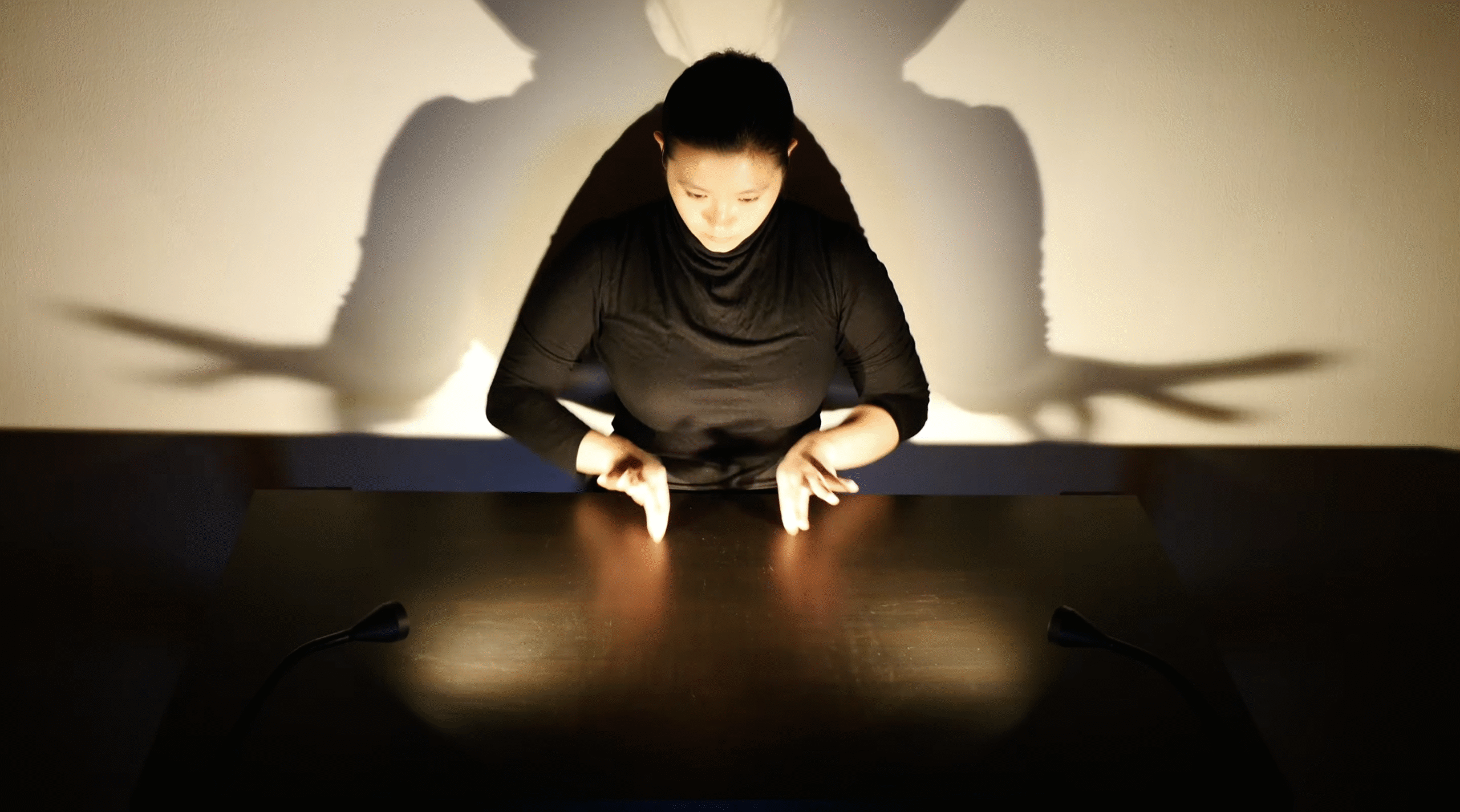 A person dressed in black sits at a table under dramatic lighting. Shadows cast on the wall create the illusion of wings.