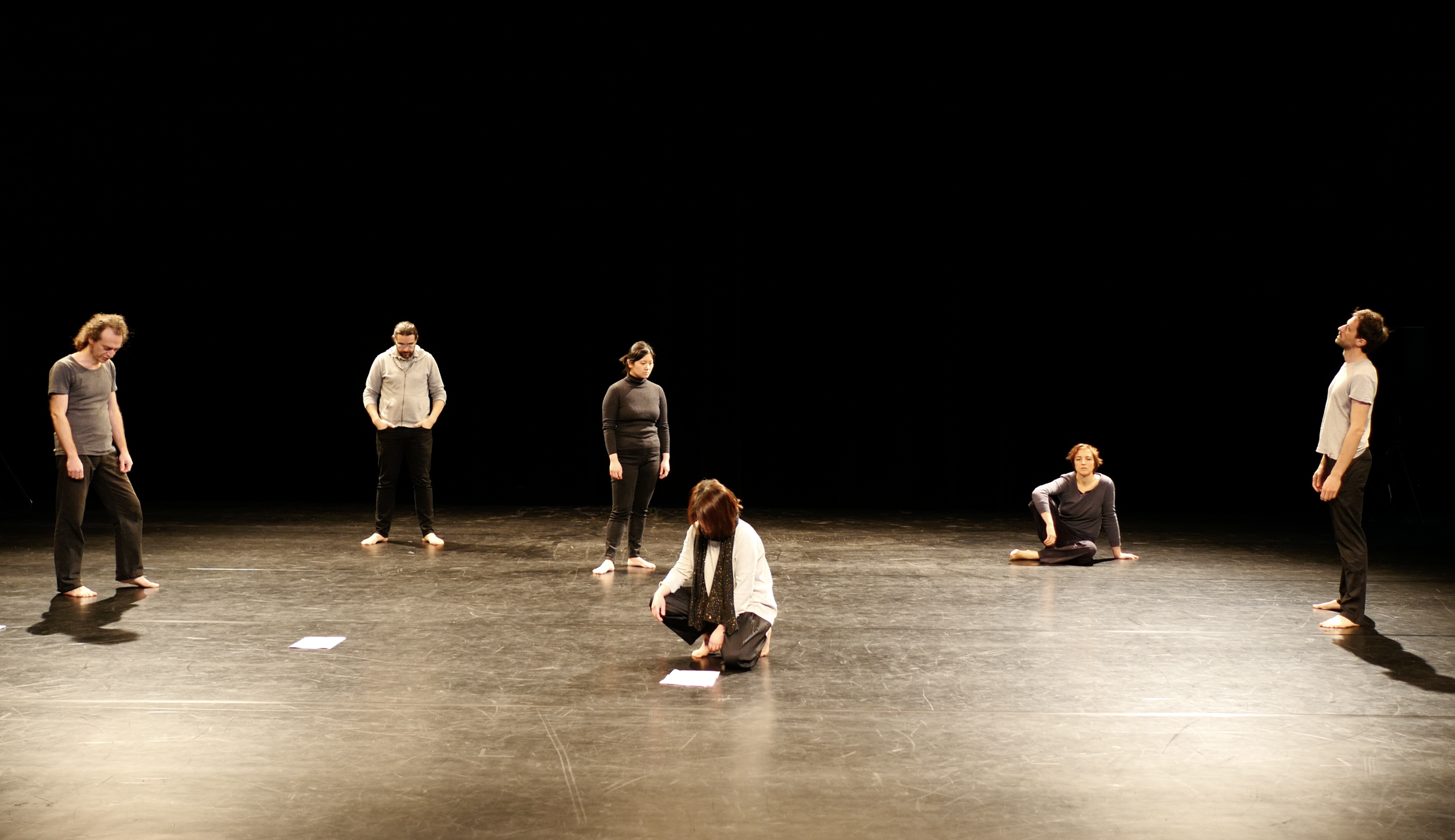 Six people are on a dimly lit stage, barefoot, in contemplative poses. Two sit on the floor, while four stand, each gazing in different directions.