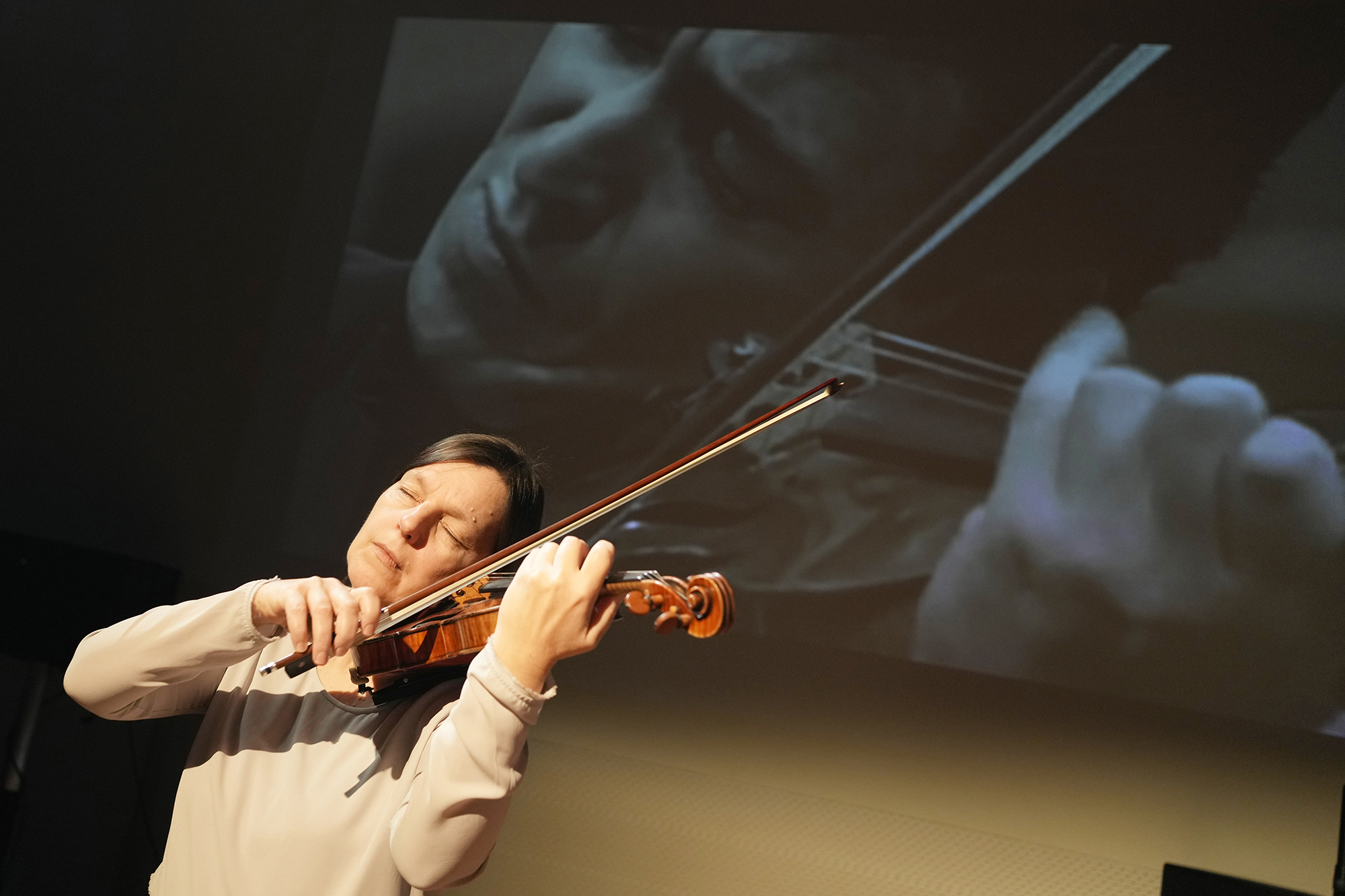 A woman passionately plays the violin with closed eyes. A large black-and-white close-up of another performance is projected in the background.