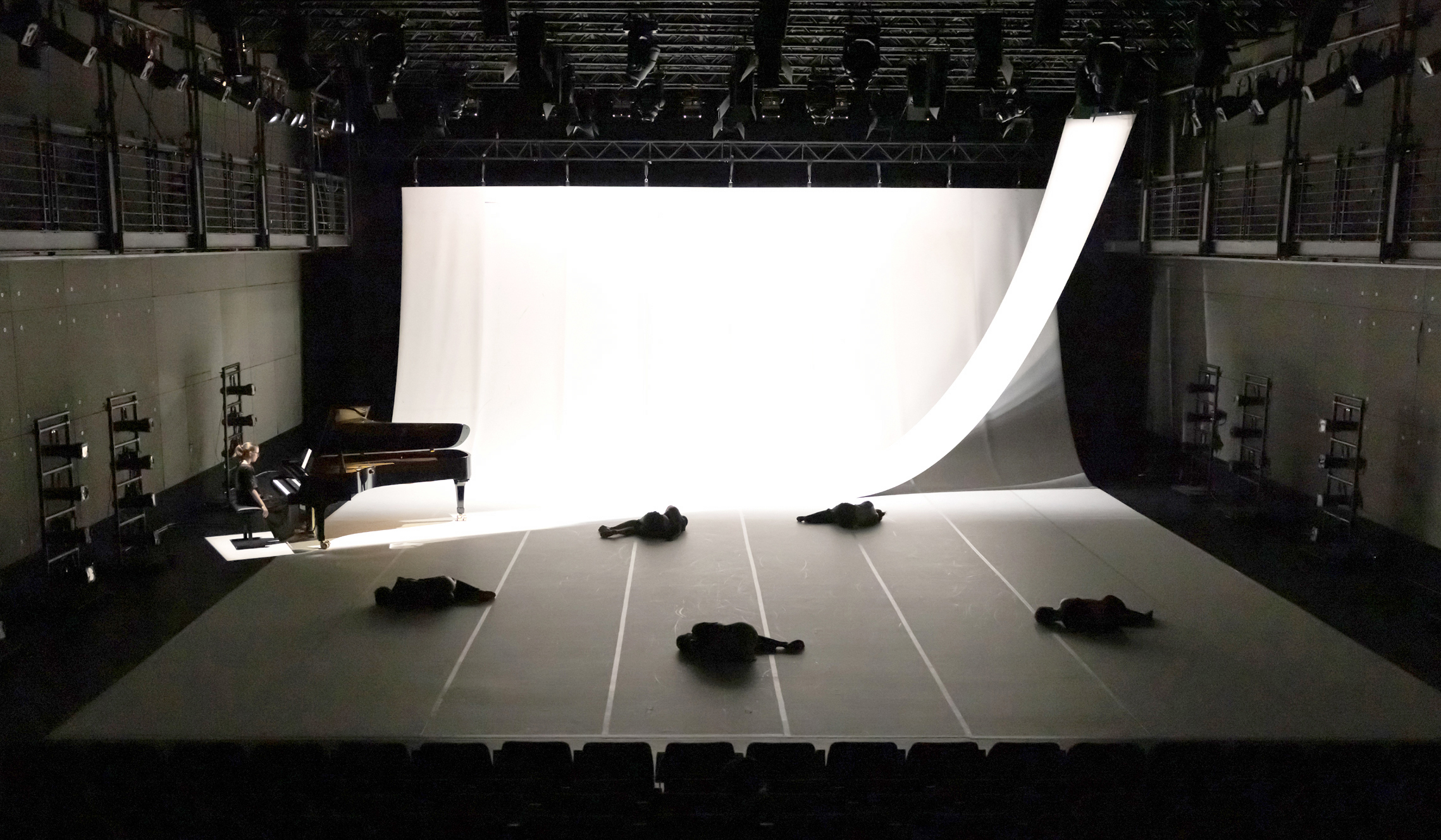 Dimly lit stage with five people lying on the floor, scattered. A pianist plays a grand piano on the left. A large white backdrop curves elegantly upward.