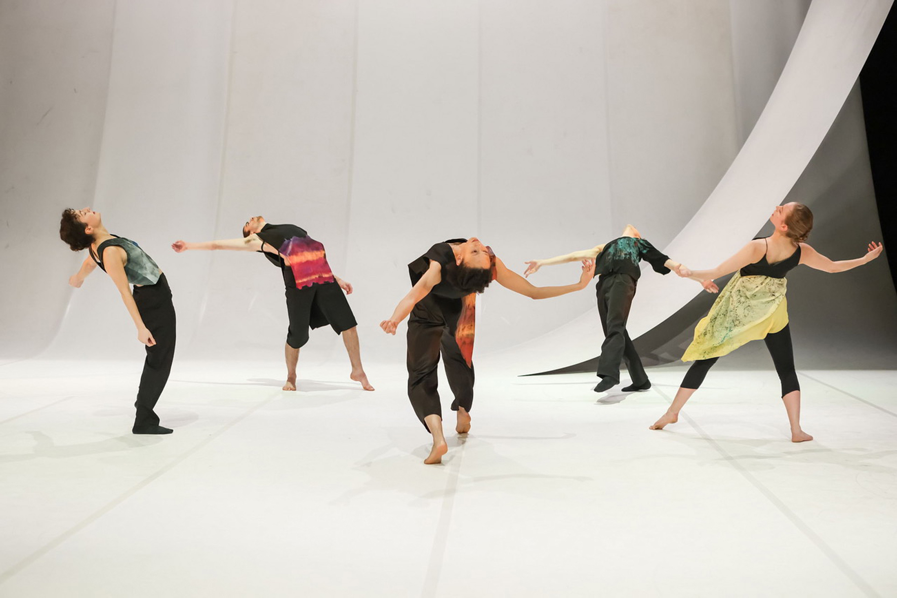 Five dancers in colorful costumes bend backward gracefully on a minimalist white stage, creating a dynamic and expressive performance scene.