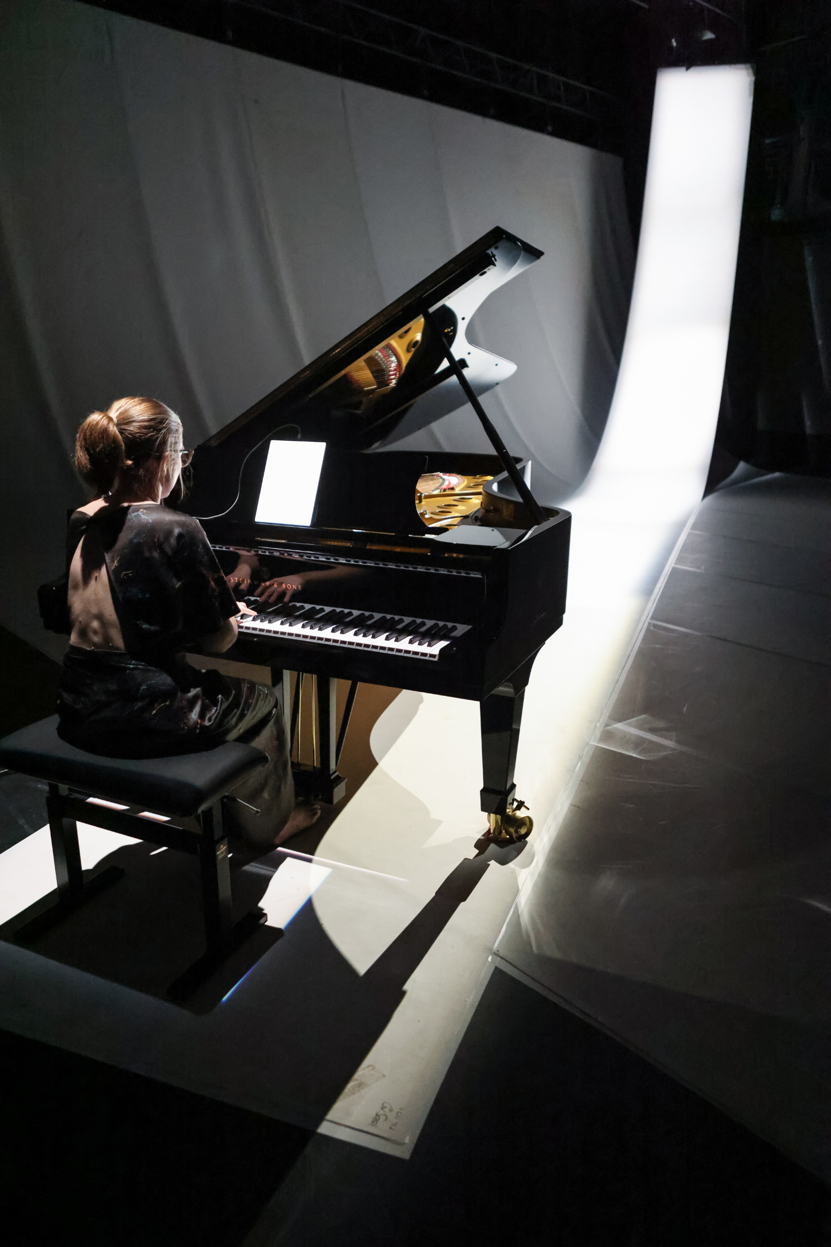 A pianist with a bun plays a grand piano on a dimly lit stage, illuminated by a soft spotlight. A long, curved white backdrop is visible behind her.