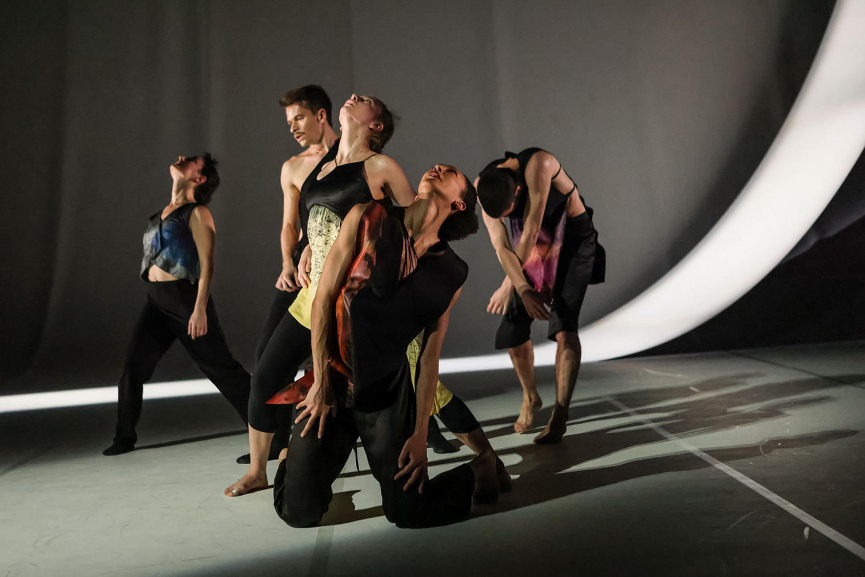Five dancers in dynamic, expressive poses on a dimly lit stage. They wear black outfits with colorful patterns.