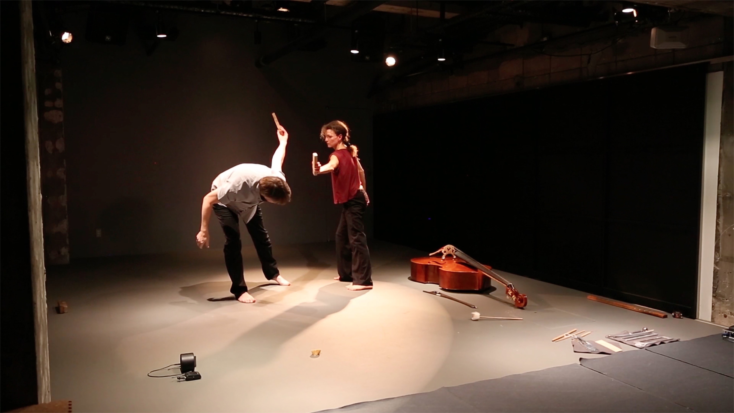 Two performers are on stage under spotlights. One is bent forward with an arm raised, and the other stands assertively pointing. A cello lies on the floor.