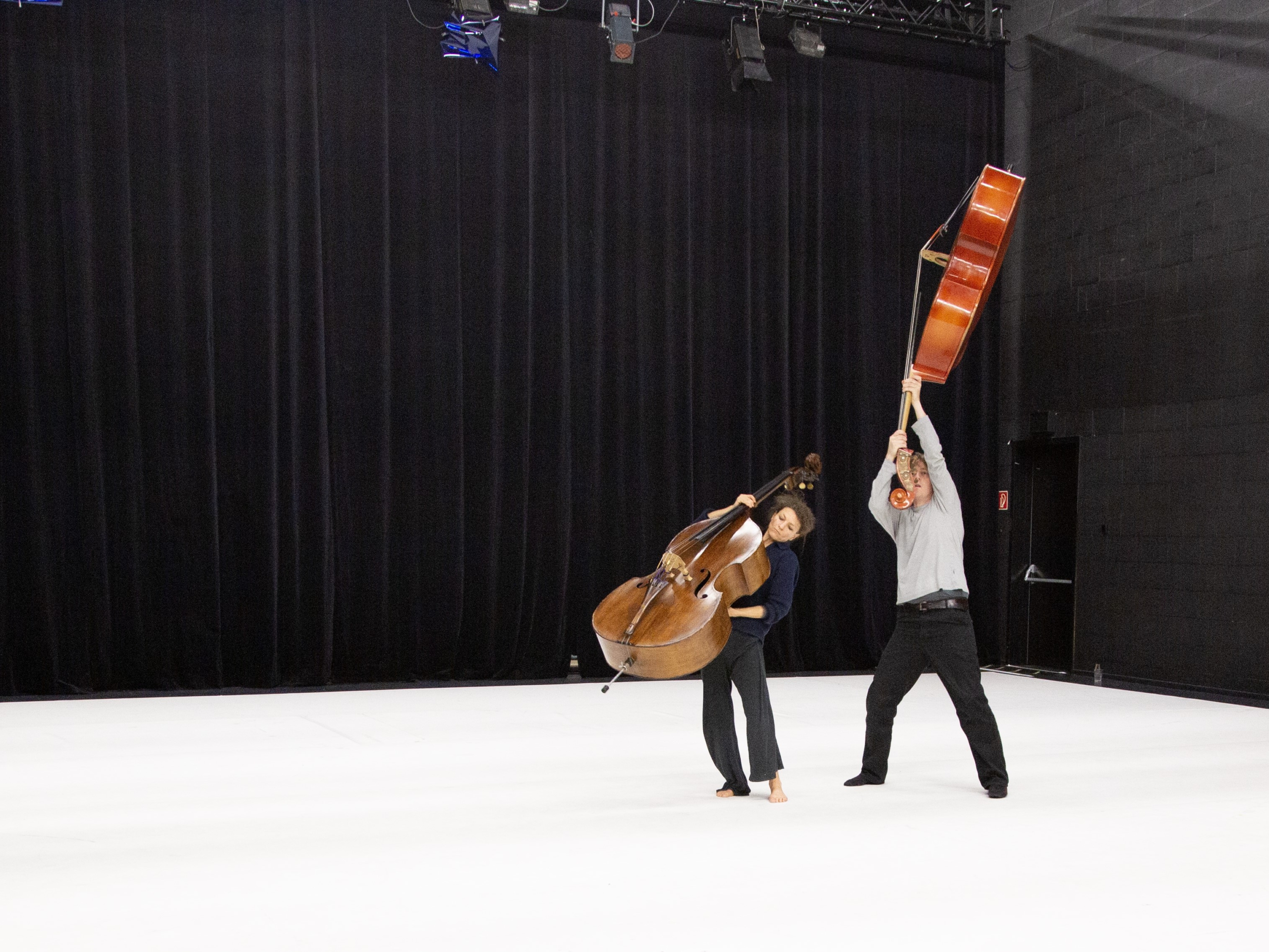 Two people perform with double basses in a spacious room. One holds the bass upright, while the other lifts theirs overhead.