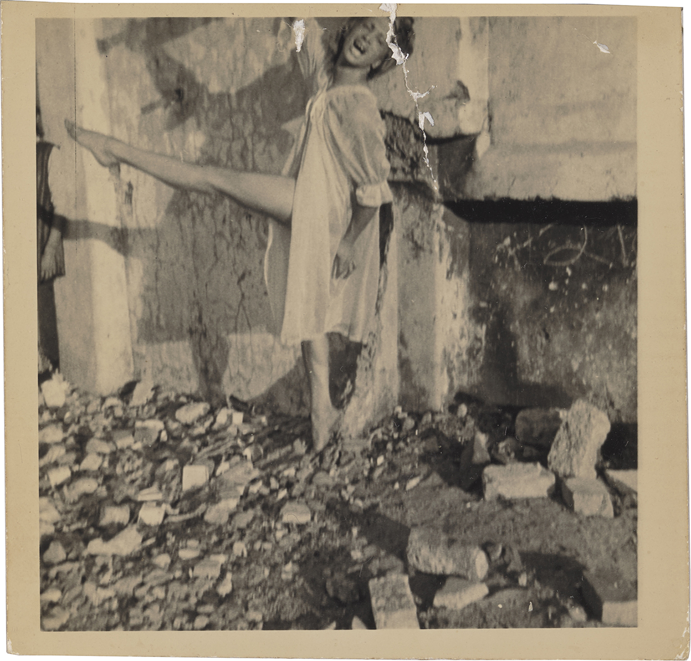 Author’s note: Dirty Dragging is based on detailed readings of the images and understands these as situated work. Accordingly, the book functions at crucial points without the supplementary captions added here; methodologically, my approach resists the notion of supposedly objective image description and the idea that accessibility can be achieved in just a few sentences.

A sepia-toned photo shows a person dancing barefoot amid rubble, wearing a white baby-doll outfit, mouth open, right arm stretched upward, and left leg raised.