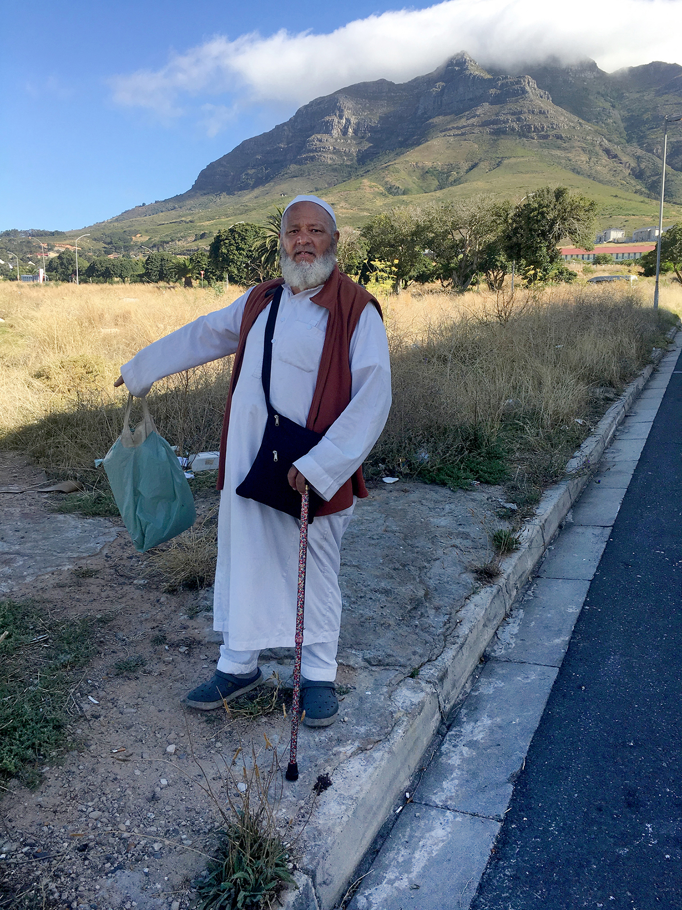 A color photo shows a person standing at the roadside before a table mountain landscape, pointing toward the ground, wearing traditional Muslim white clothing (thawb).
