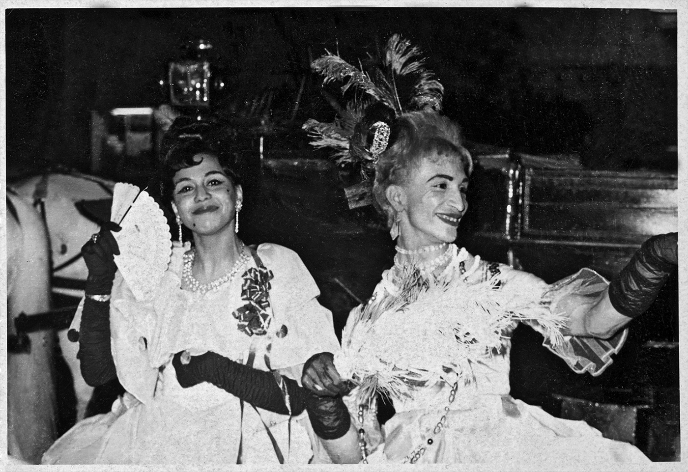 A black-and-white photo shows two laughing people in white ball gowns with feather decorations, long black gloves, and a fan, sitting in a carriage.