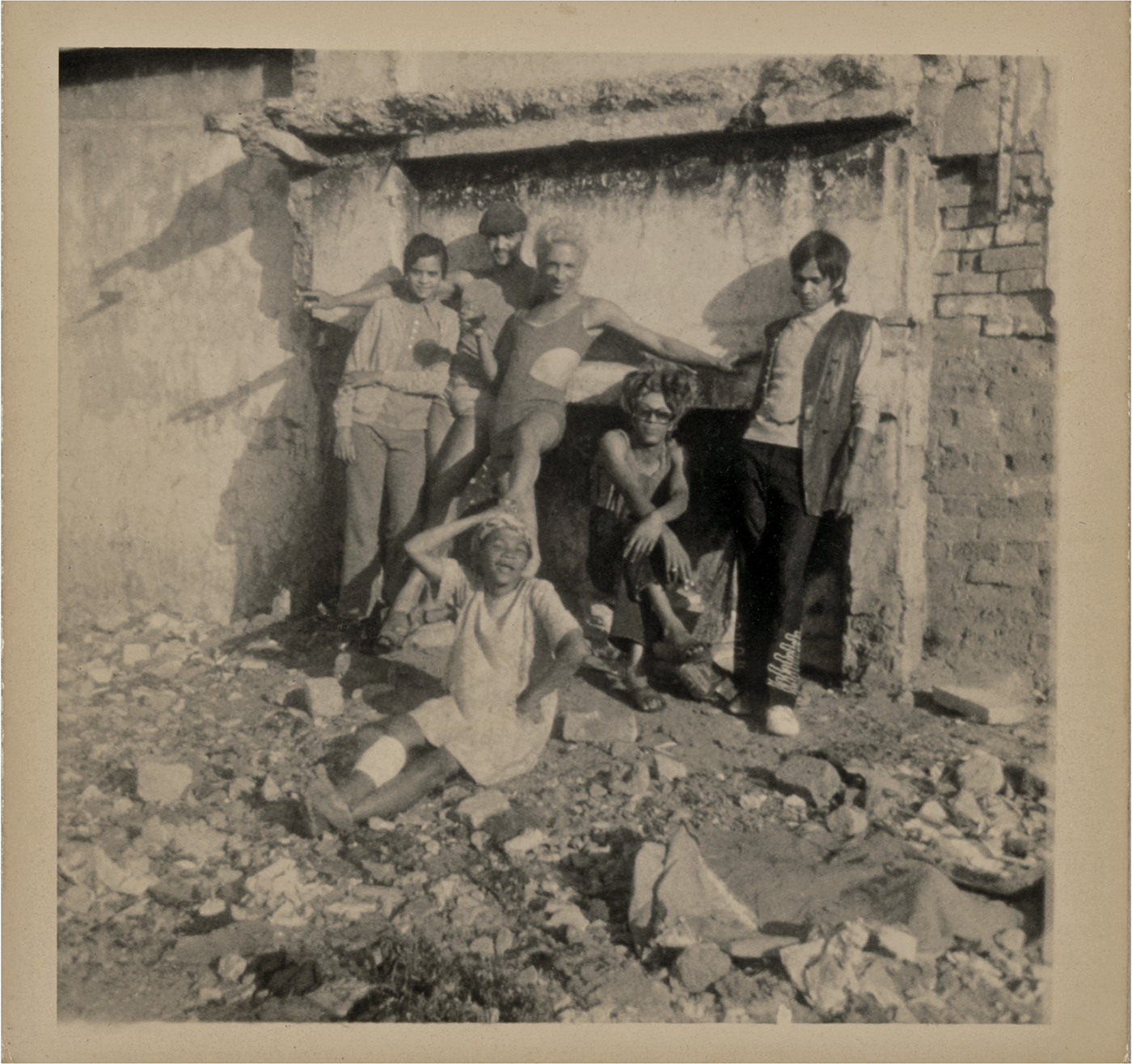 A sepia-toned photo shows six people posing amid rubble in front of a wall.