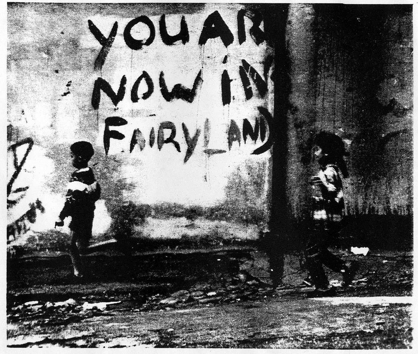 Ein Schwarzweißfoto zeigt ein Graffitti an einer Hauswand („You are now in Fairyland“), an der zwei Kinder vorbeigehen.