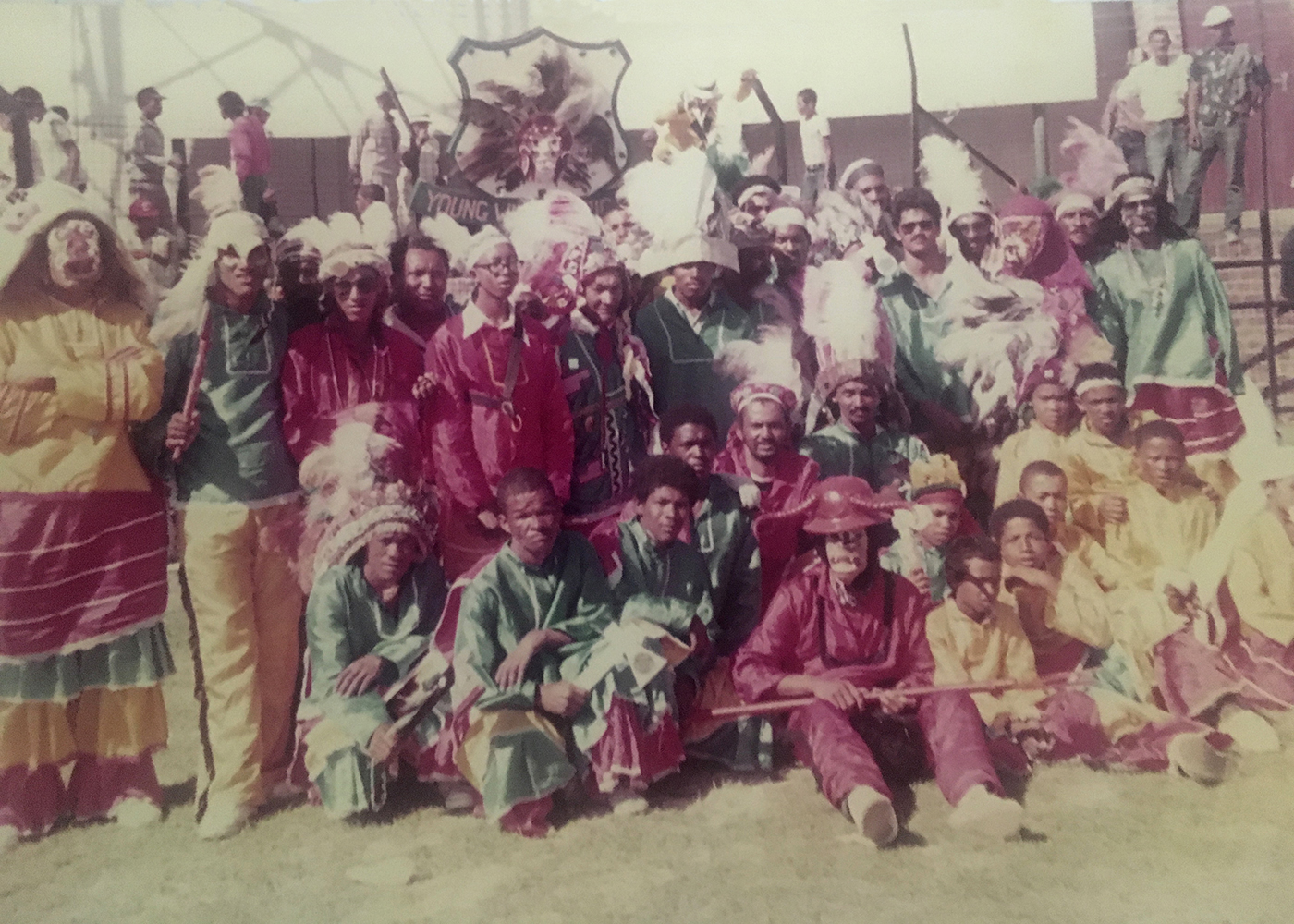 Ein Farbfoto zeigt eine farbig kostümierte Gruppe mit Federschmuck und Gesichtsmasken.