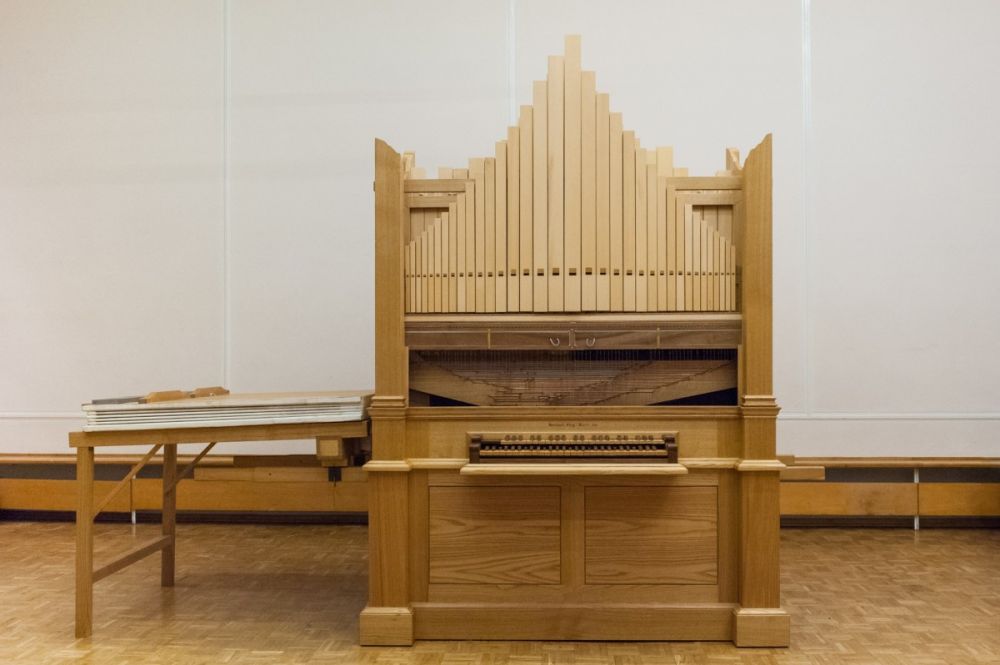 A photograph of a wooden pipe organ with a single manual (keyboard) and vertical pipes arranged symmetrically. The instrument is placed in a room with wooden flooring and white walls, and a table with the bellows is positioned to its left.