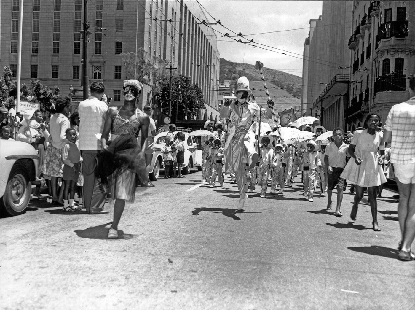 Ein Schwarzweißfoto zeigt einen Karnevalsumzug mit einer Drag Queen, einem Vortänzer und einer Gruppe an Kindern in hellen Kostümen, mit Sonnenschirmen und in Blackface, die auf der Straße tanzen, während Zuschauende am Straßenrand stehen.