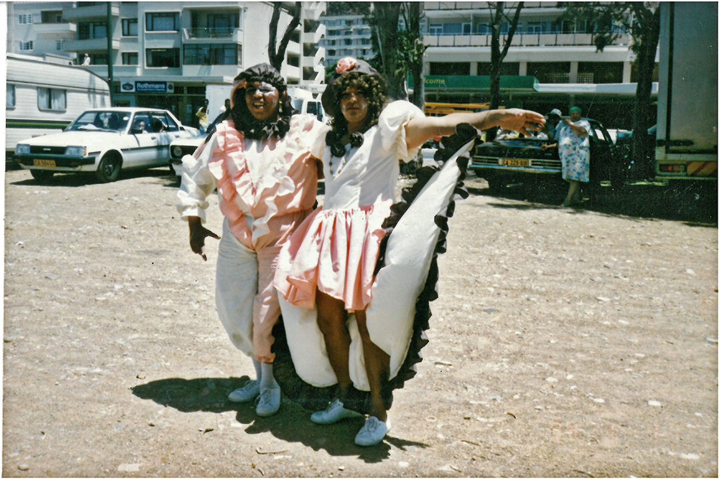 Ein Farbfoto zeigt zwei Personen, die auf einem Parkplatz in weiß- und rosafarbenen Kostümen für die Kamera posieren.
